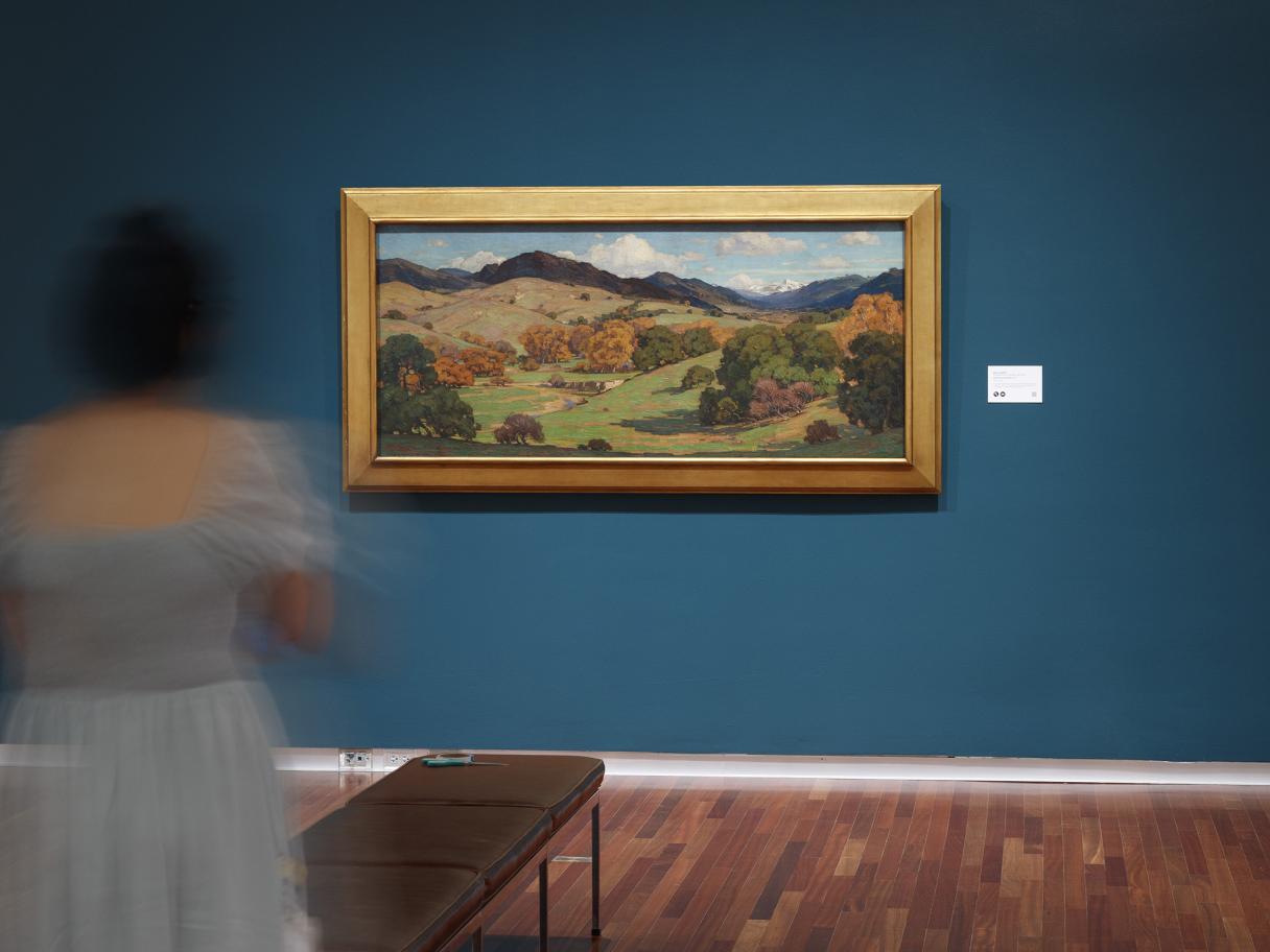 A person with dark hair in a light blue dress stands behind a bench, facing a gold-framed landscape painting.