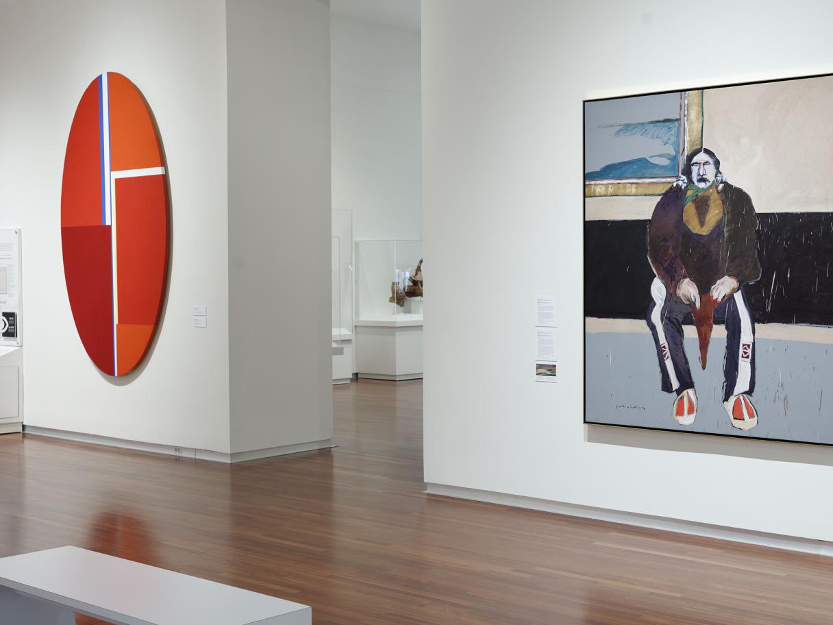 A large red oval hangs on a white wall next to a painting of a Native American man sitting.