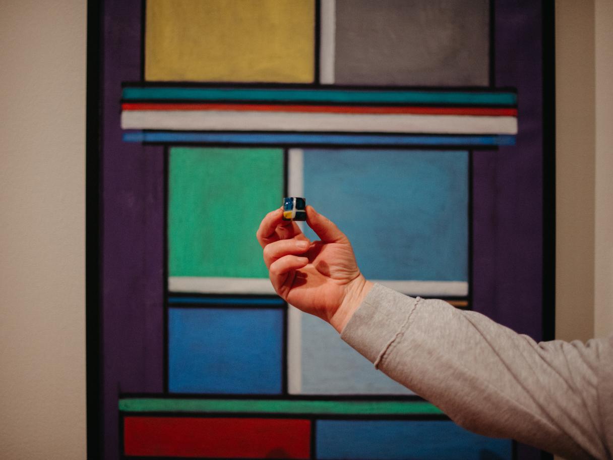 A hand holds a colorful chocolate in front of a color-block painting of the same pattern.