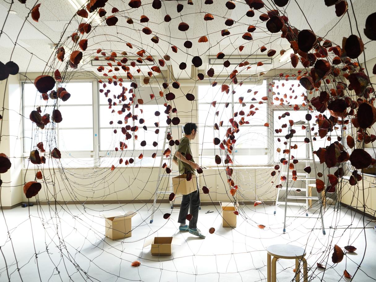 A man stands in the middle of a room with boxes, stools and ladder and small rock shapes copper pieces hanging from wire spread across the room.