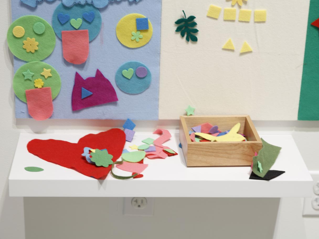 A table covered in felt shapes. Behind it there is a blue and white felt board with colorful felt shapes arranged on it.