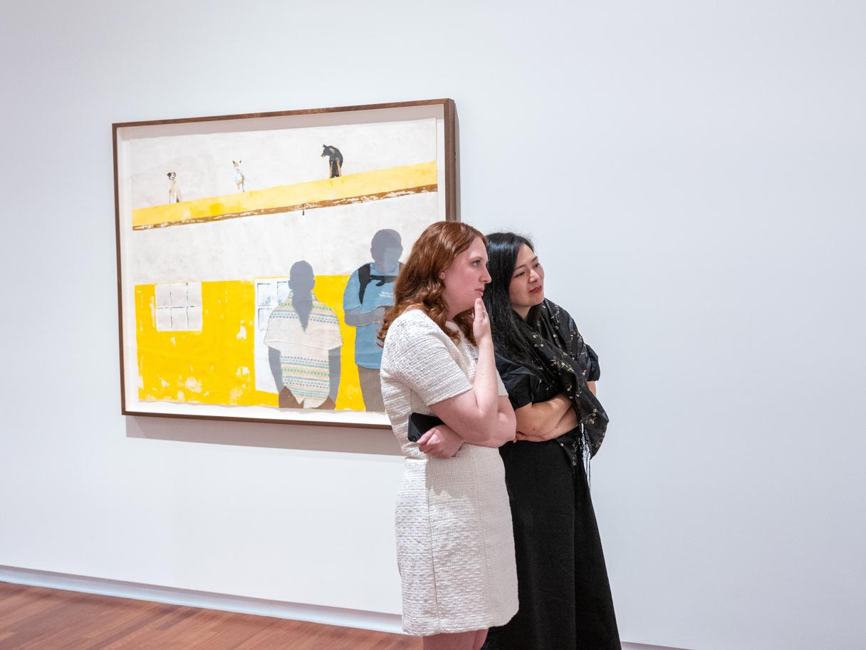 salt 16: Arleene Correa Valencia Two women in dresses stand looking at art off view. There's a framed yellow artwork depicting two human silhouettes hanging on the wall in the background..