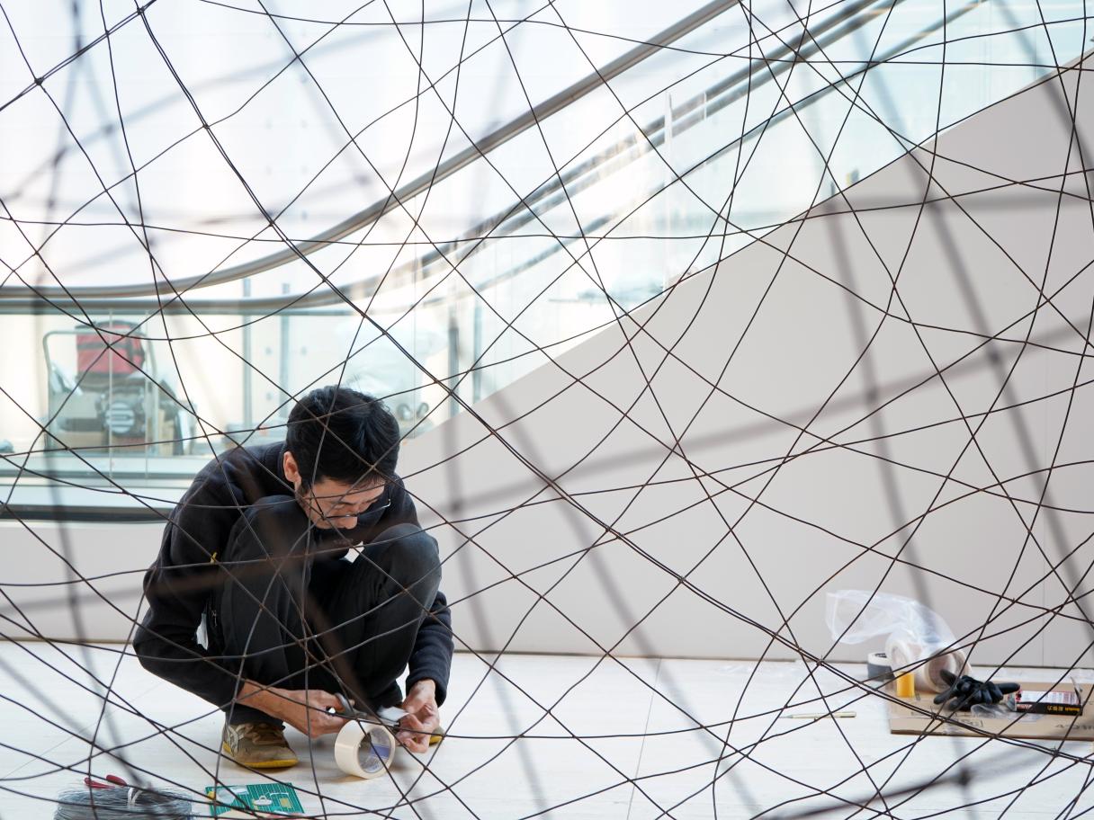 Artist, Minako Yoshida kneels down to work. He is surrounded by wire.