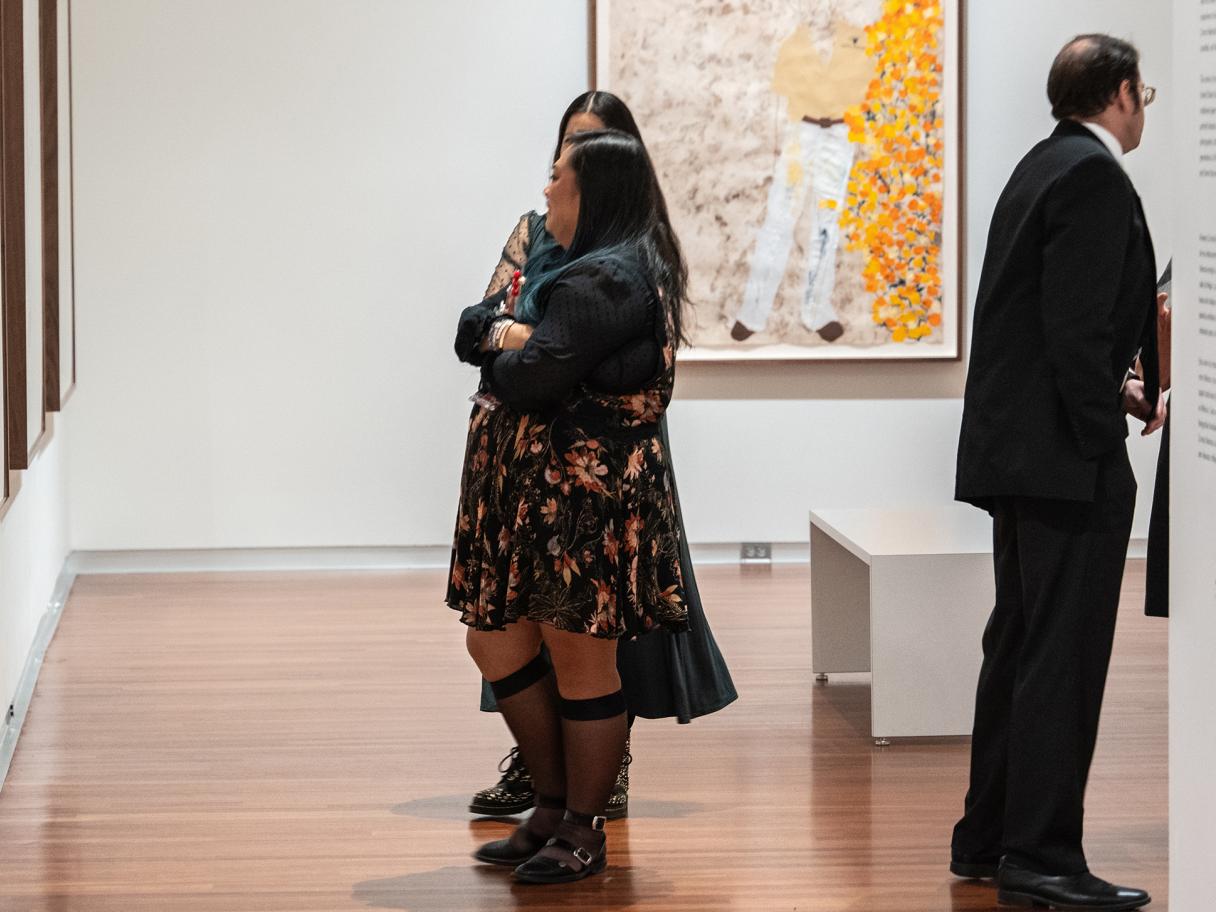 Two women with long dark hair stand next to one another, speaking and looking at art hung on a wall.