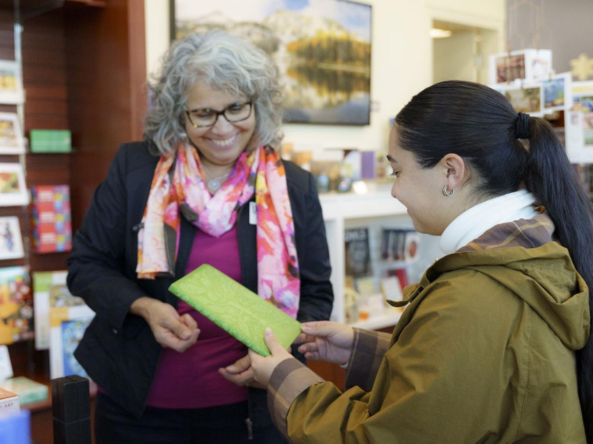 Woman with long dark hair, pulled back, wearing a coat holds a book given to her by another woman in a pink floral scarf with curly gray hair.