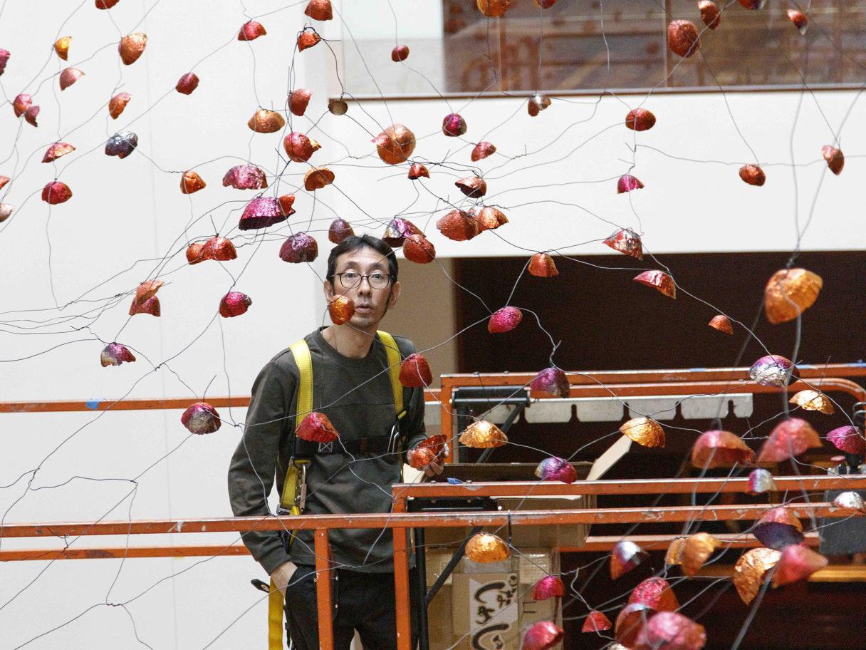 A man stands on a lift in the middle of an art installation of wires and foil, copper cups.