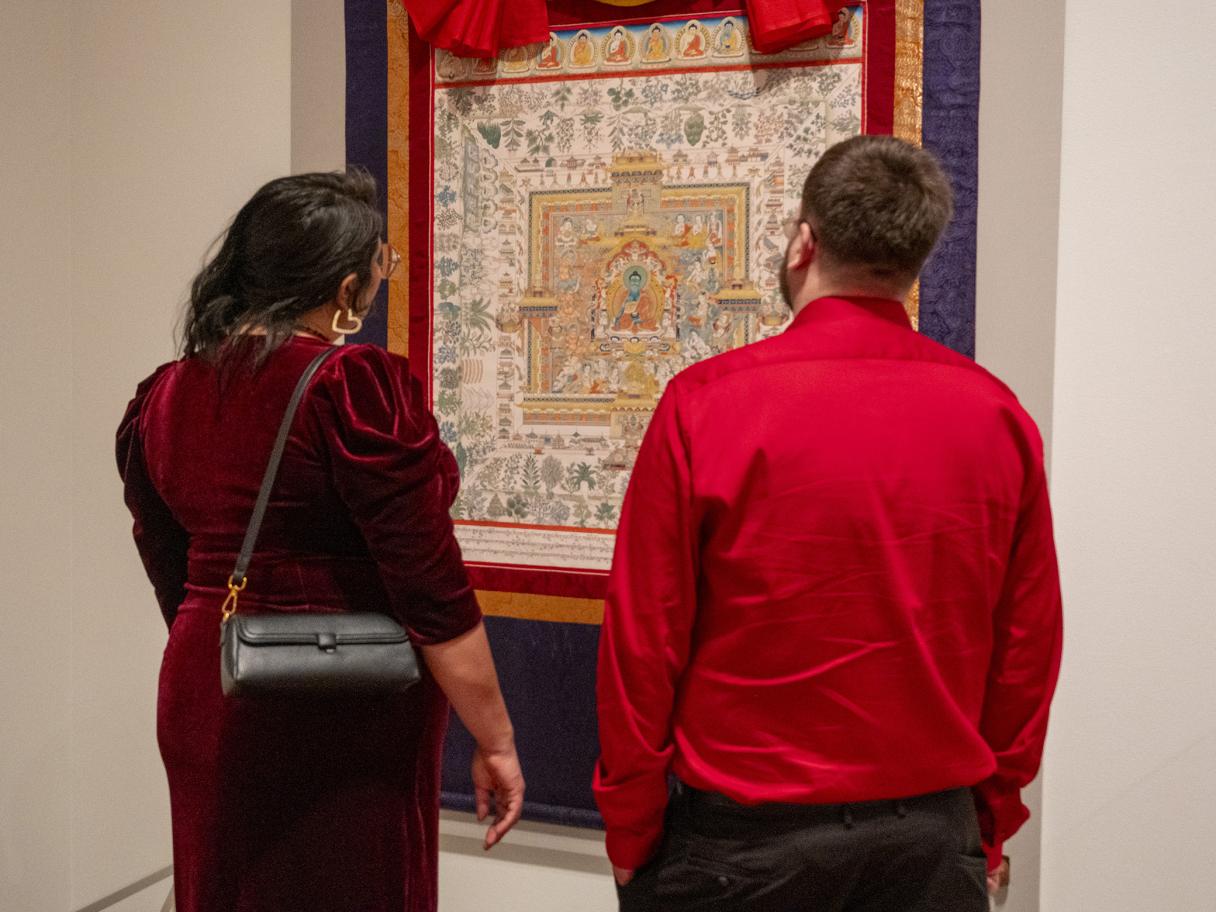 A woman in a long red dress and a man in a long sleeve shirt stand together looking at a Himalayan Art on the wall.