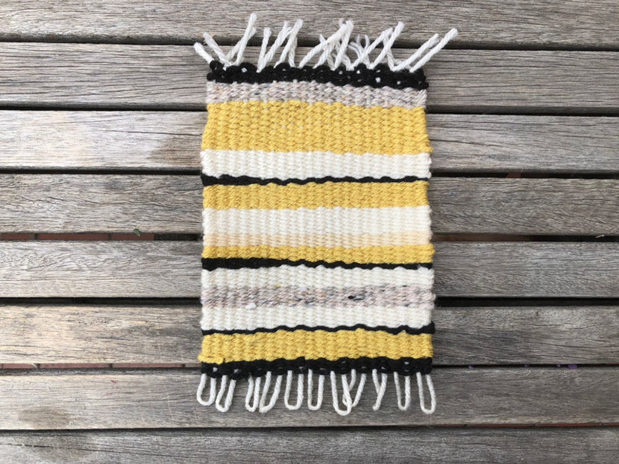 A small woven rug on a wooden background. The rug has black, yellow, beige, and white stripes of different thicknesses.