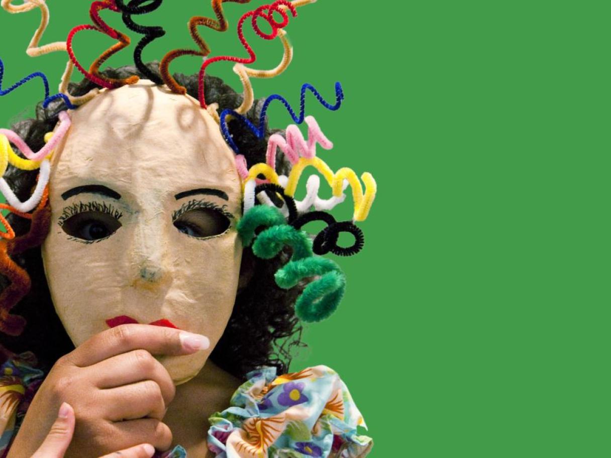 A child with a mask over their face. The mask has curly hair made of colorful pipe-cleaners.