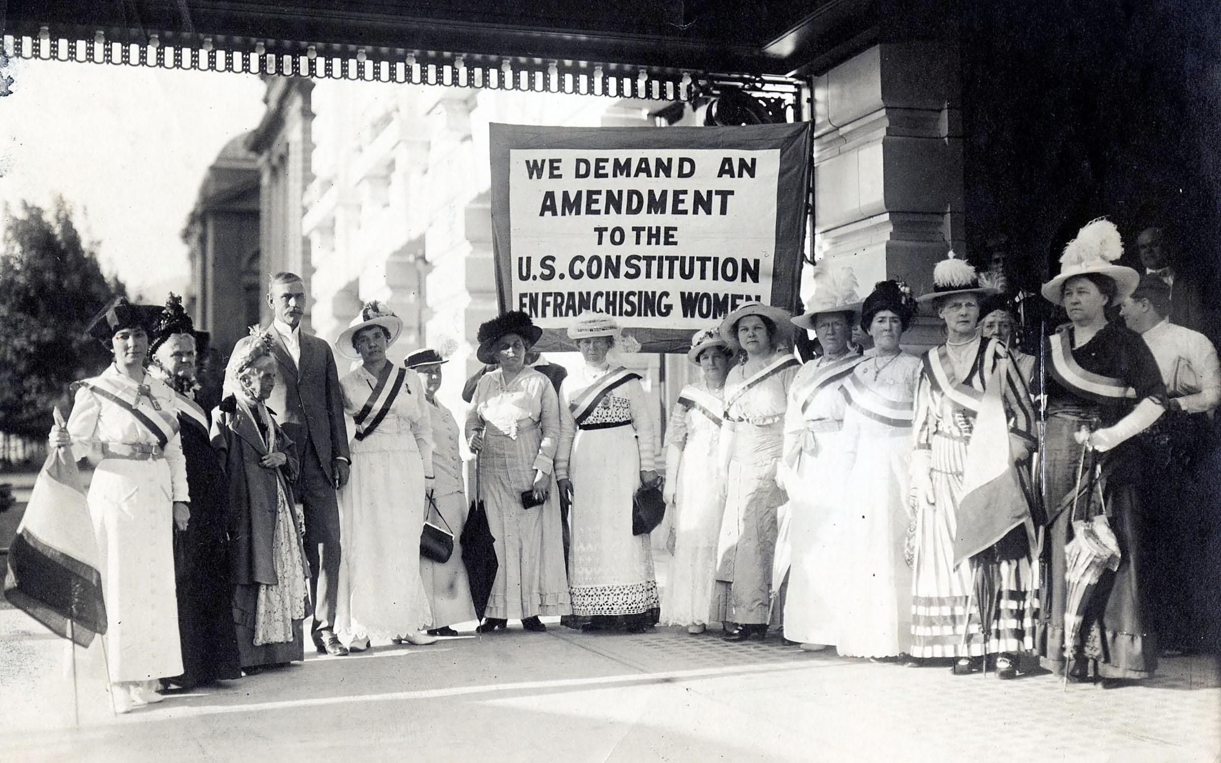 Susan B. Anthony stands with a group of  Women Suffrage Leaders in a group in front of Hotel Utah in 1915 behind the groups a banner reads We demand an amendment to the US Constitution Enfranchising Women. 