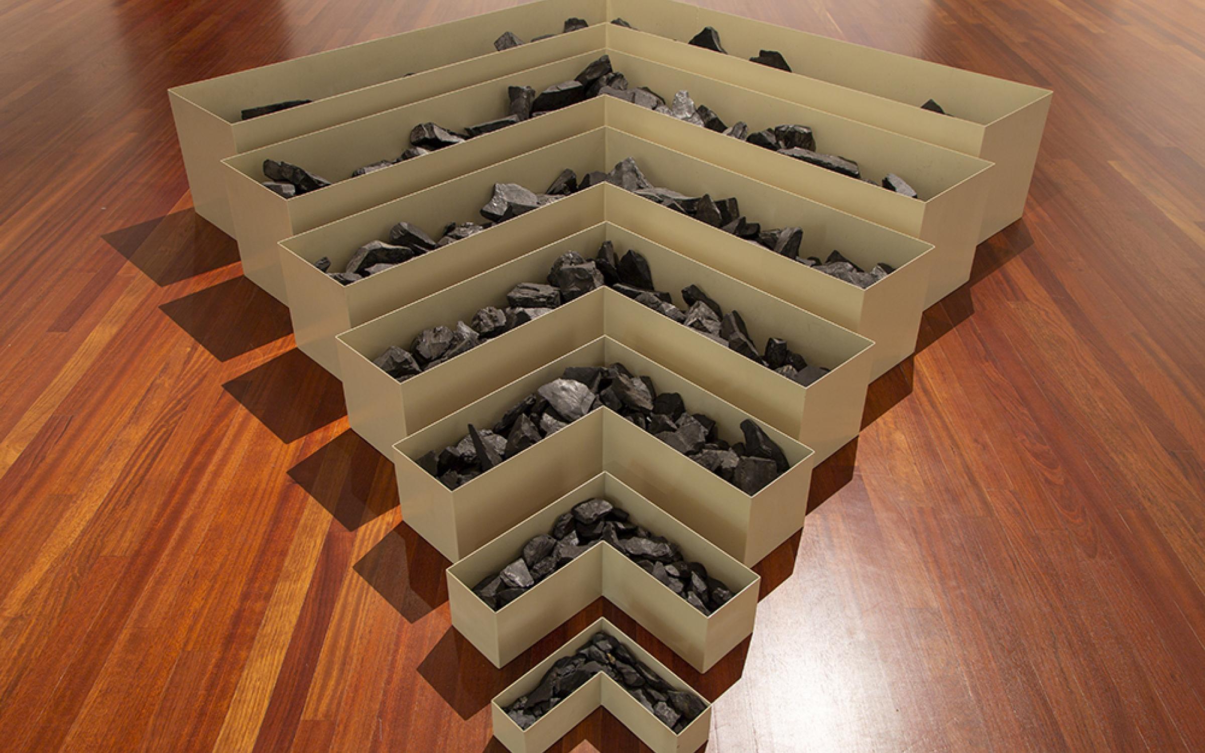 A set of 7 nested V-shaped cream colored metal boxes are lined up from small to large forming a tiered shaped meant to resemble a strip mine. They are filled with coal. Robert Smithson, Nonsite, Site Uncertain, 1968 