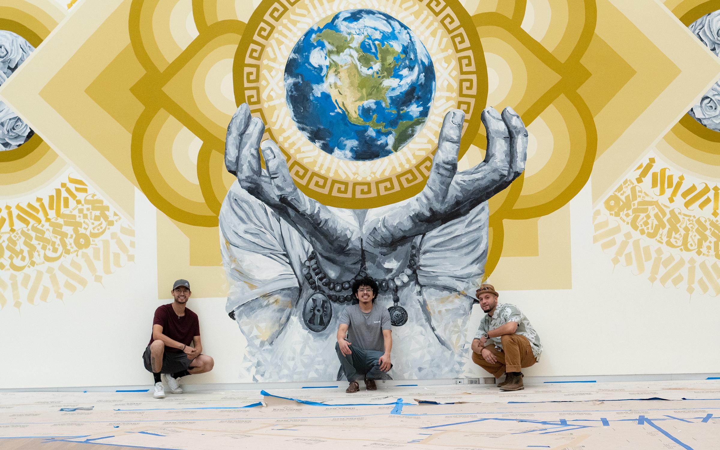 2020 from here on out in process Three artists kneel in front of a mural depicting a persons hands holding the Earth surrounded by a geometric mandala in golden hues.