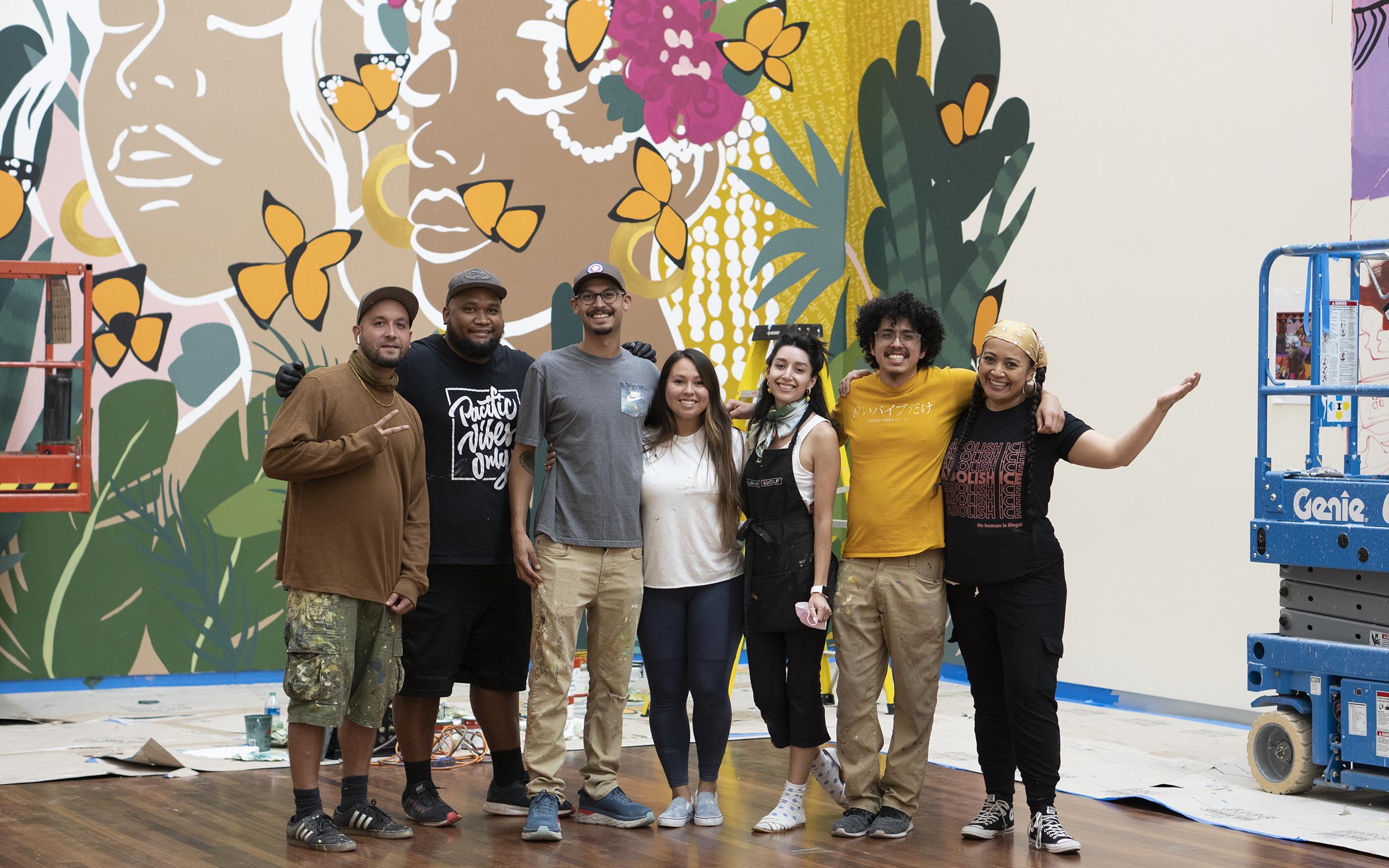 The seven muralists of 2020: From here on out posing infront of a mural in the UMFA Great Hall 