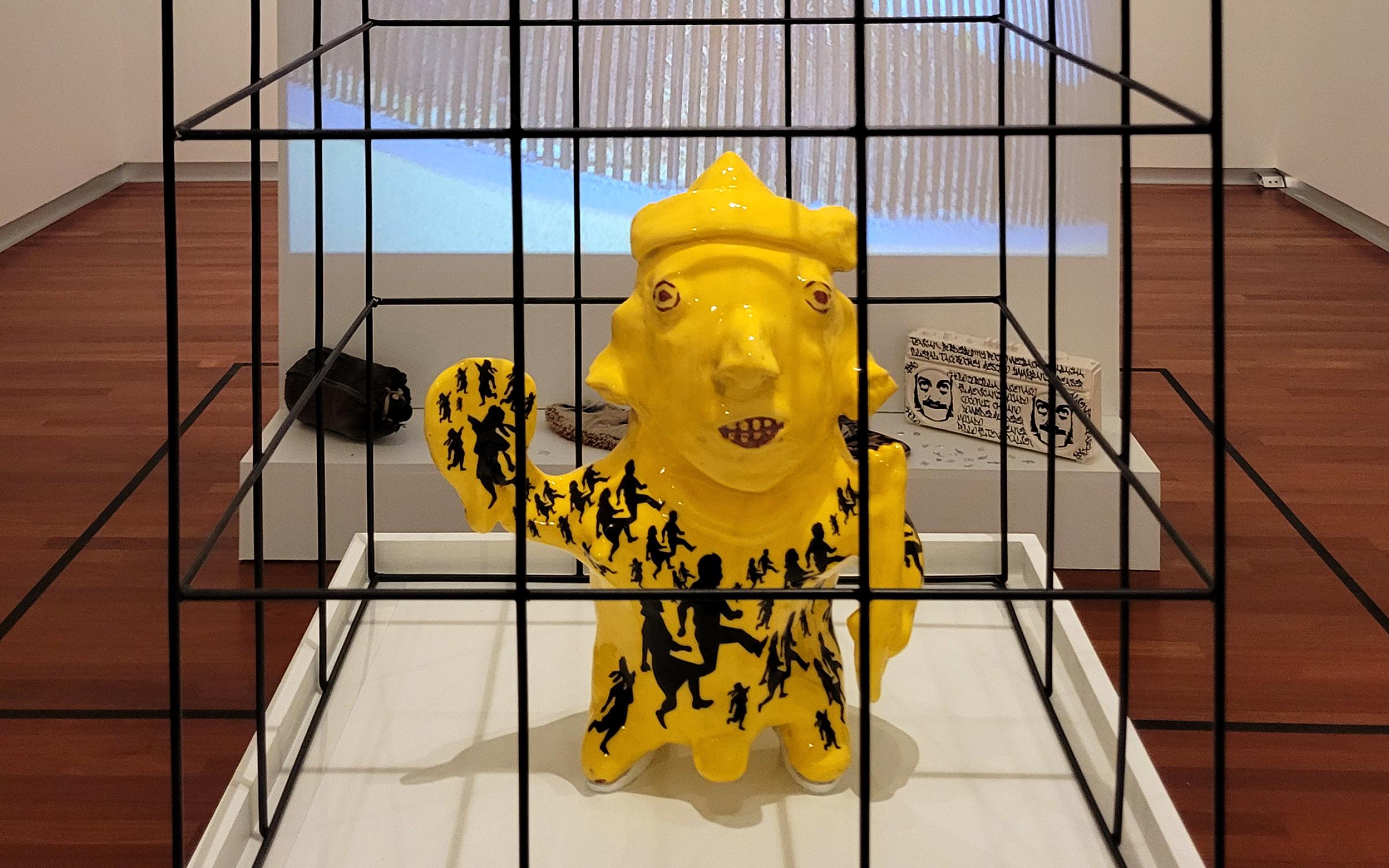 a replica of an ancient mesoamerican sculpture of a child glazed bright yellow with a pattern of silhouetted children across the body, the sculoture sits in a black cage 