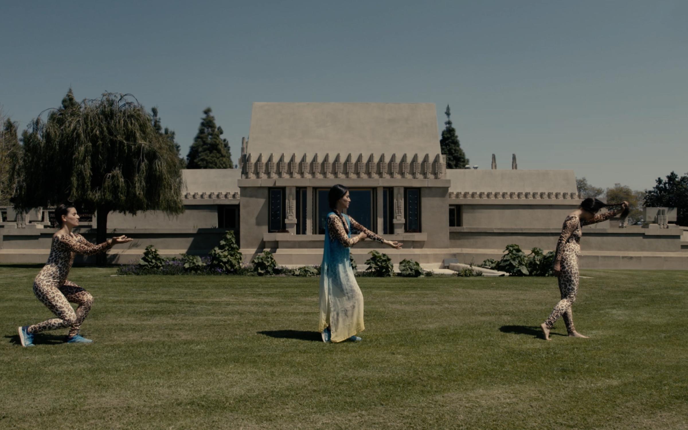 Three women dancing through a grass lawn in front fo a gray, cement building