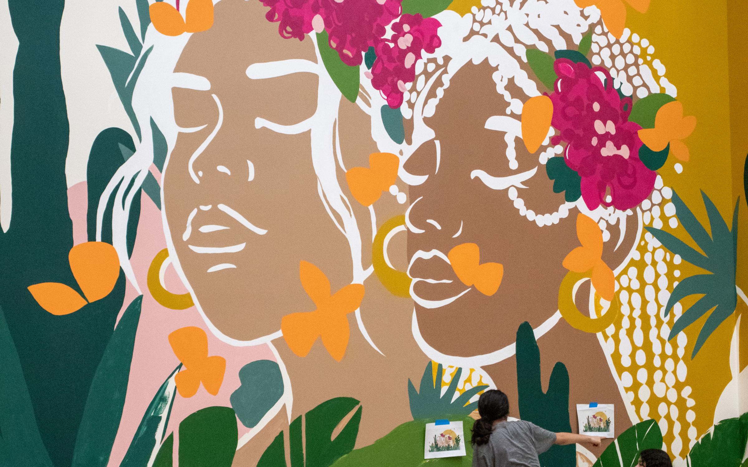 Young women painting a mural of two brown women with pink flowers in their hair.