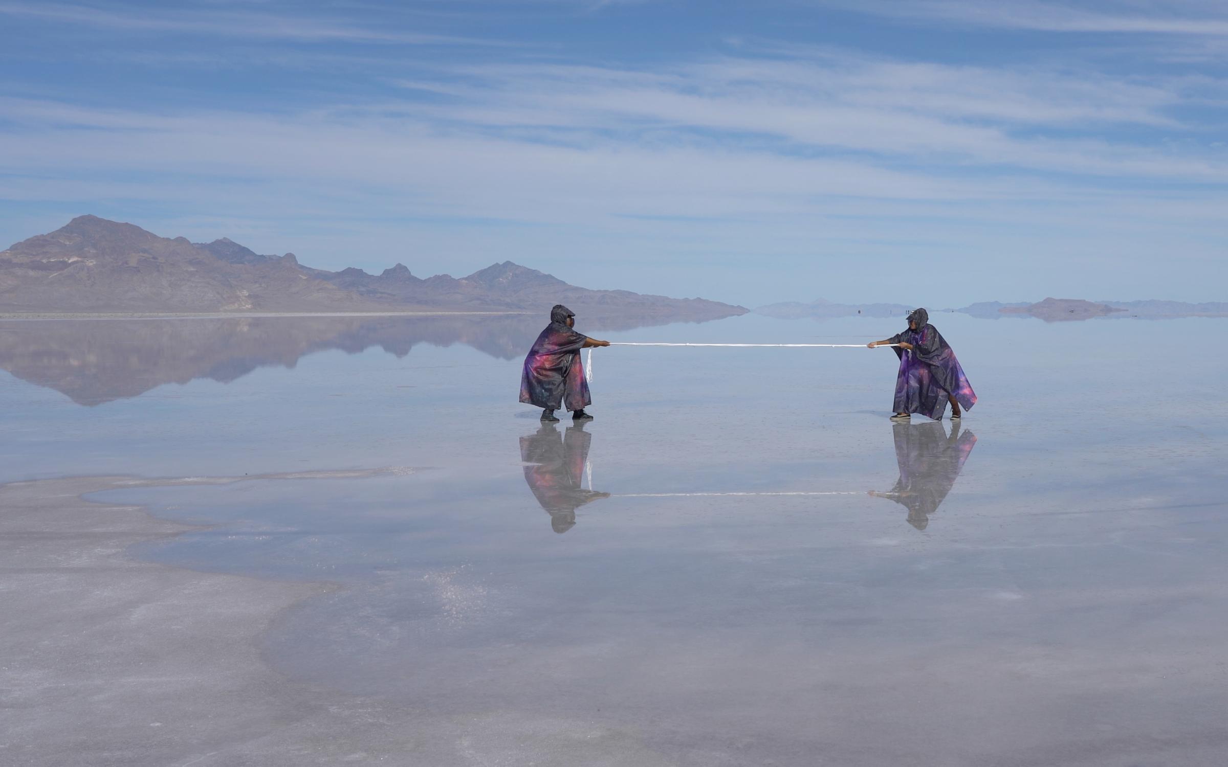 Two black cloaked people pulling a string in a reflective lake with a blue sky and shadowy mountain.