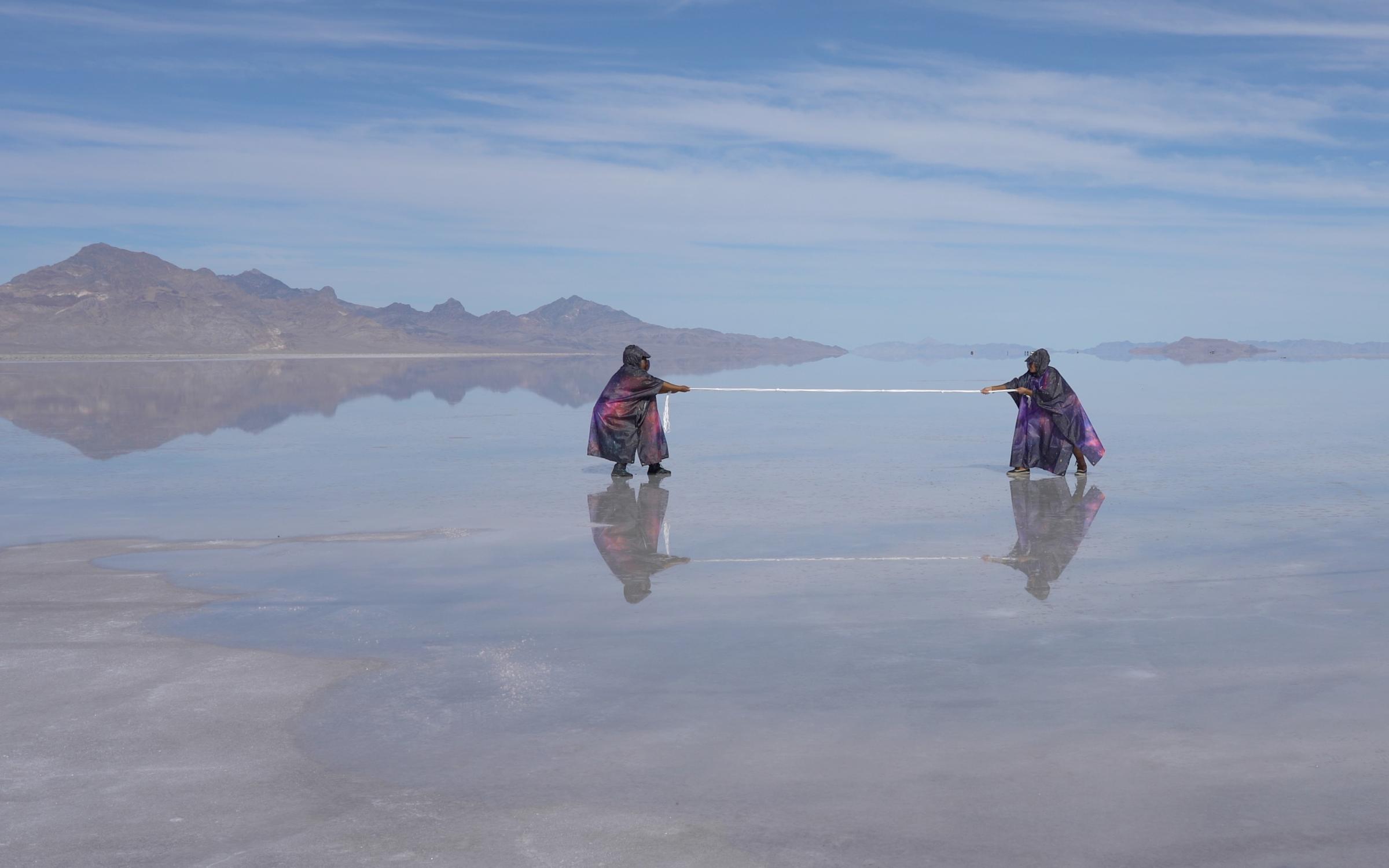 D.R.E.A.M = AFRAM by Alisha B. Wormsley Two black cloaked people pulling a string in a reflective lake with a blue sky and shadowy mountain.