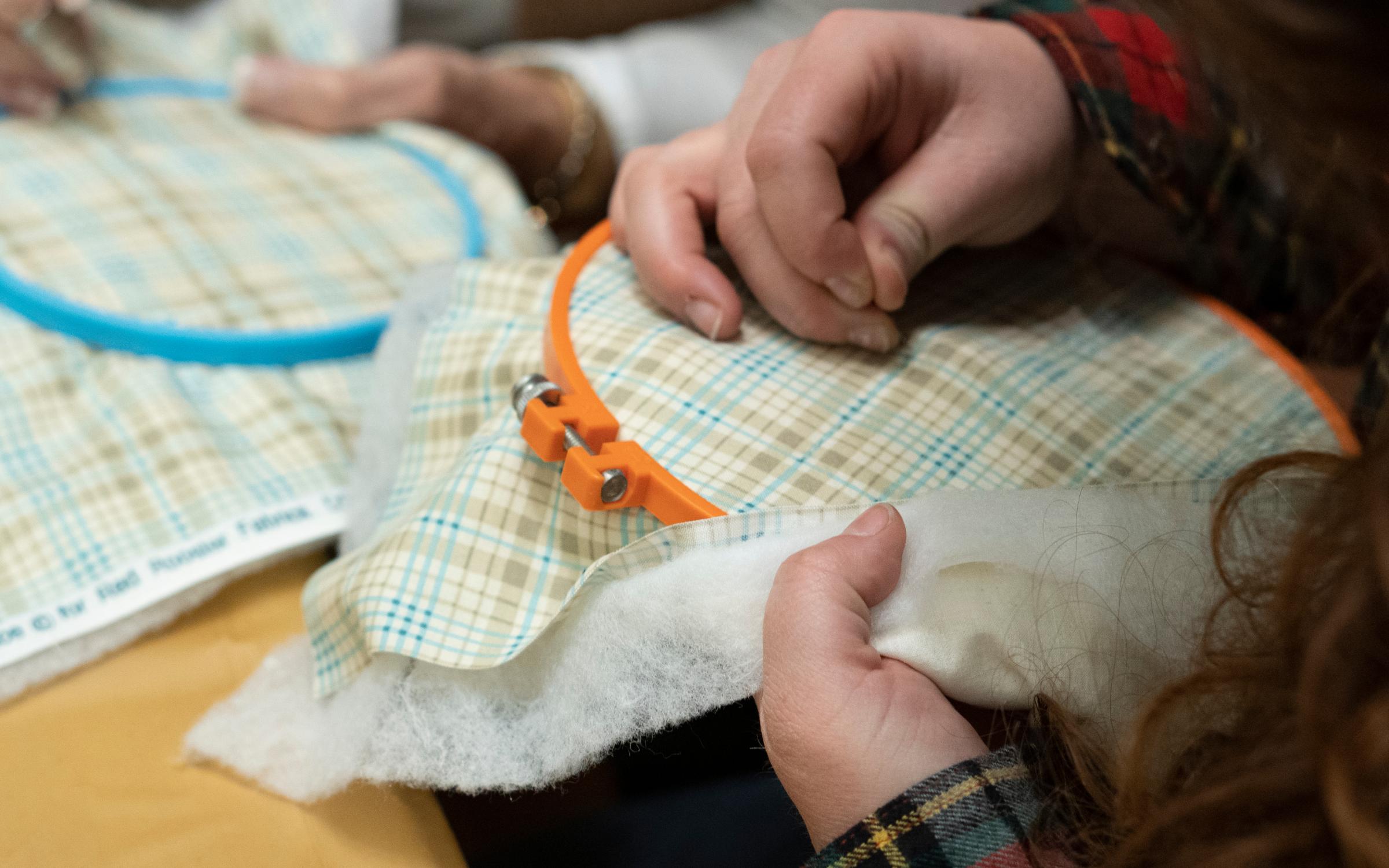 a close up image of a person's hands holding an orange embroidery hoop with plaid fabric in one hand and a needle stitching in the other