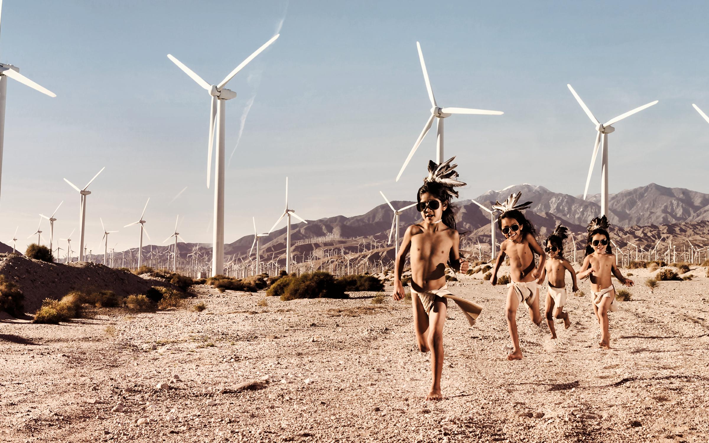 A group of three Indigenous boys in traditional garb running on a desert landscape with wind turbines in the background.