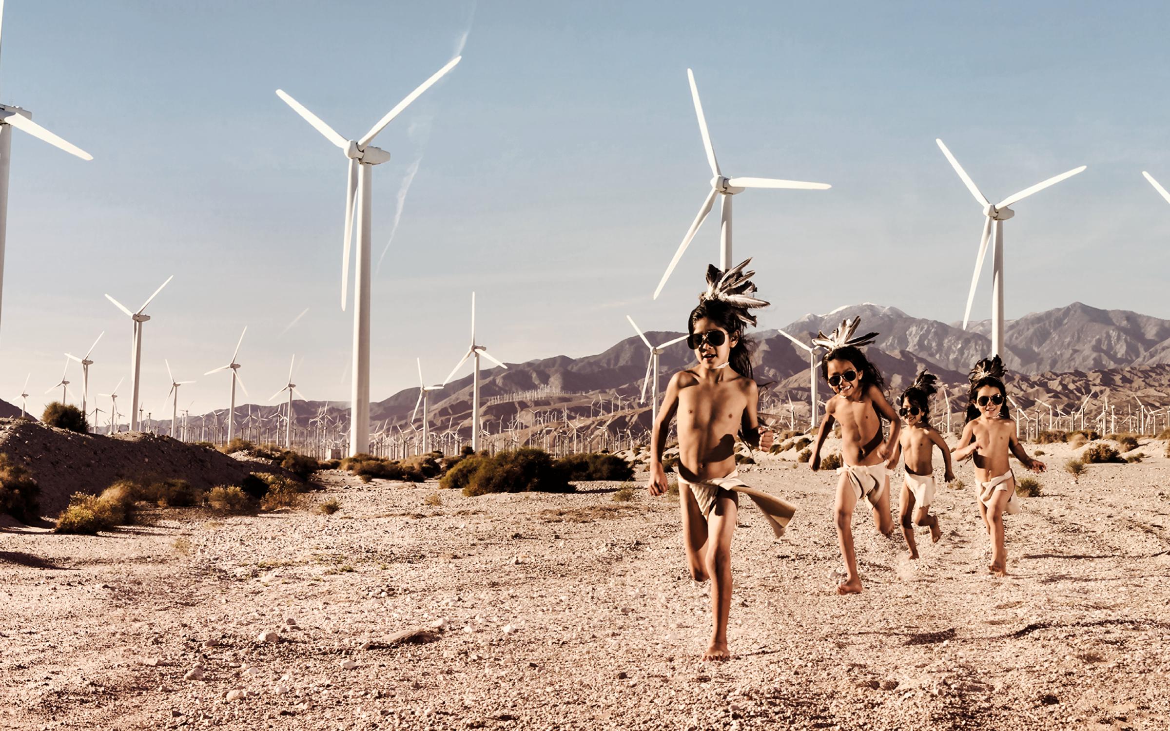 Cara Romero (Chemehuevi, born 1977, lives Santa Fe, NM), Evolvers, 2019, archival pigment print, purchased with funds from the Paul L. and Phyllis C. Wattis Fund, UMFA2022.3.1 A group of three Indigenous boys in traditional garb running on a desert landscape with wind turbines in the background.