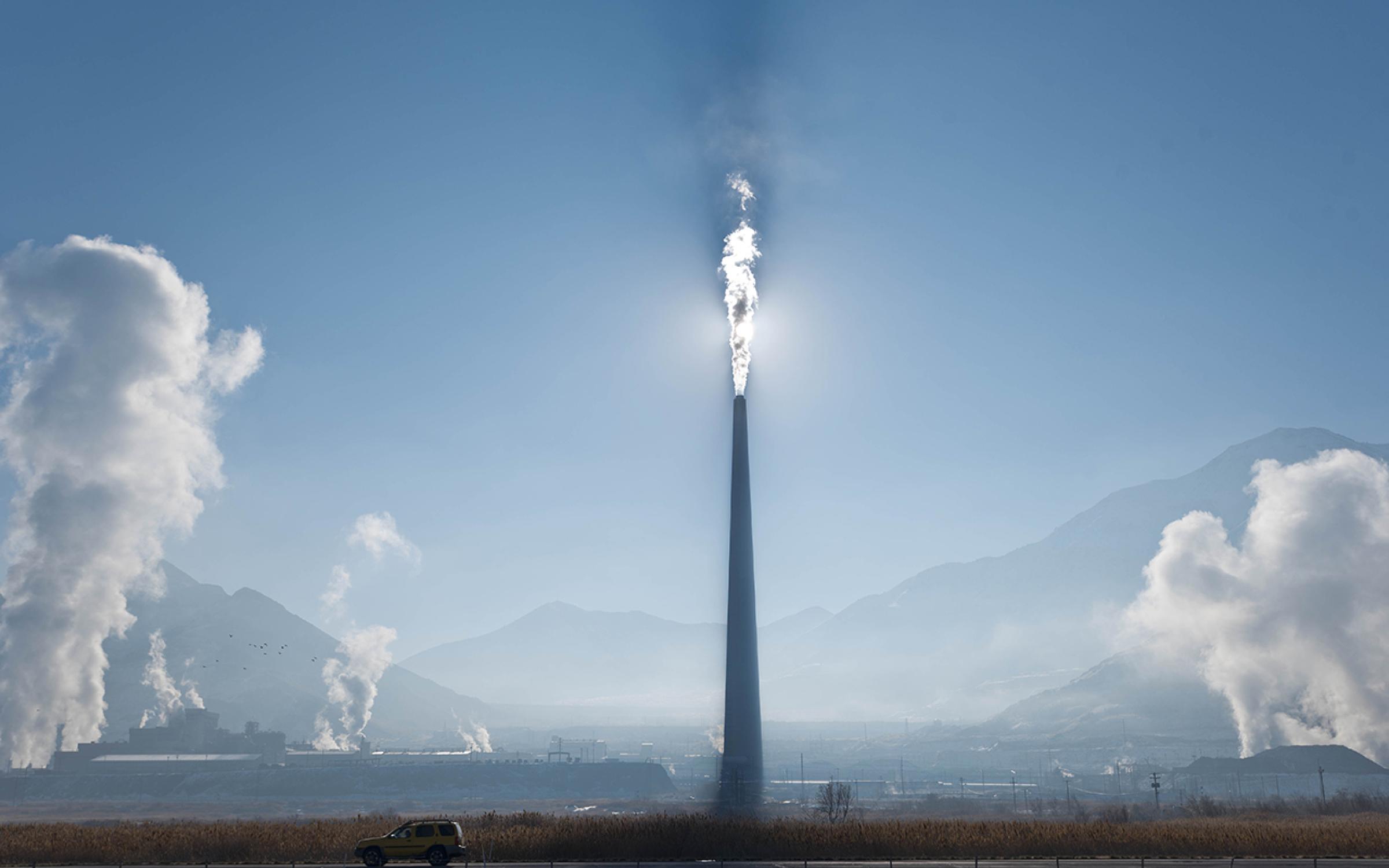 A large smelter with white smoke coming out of it in front of the sun in a blue sky.