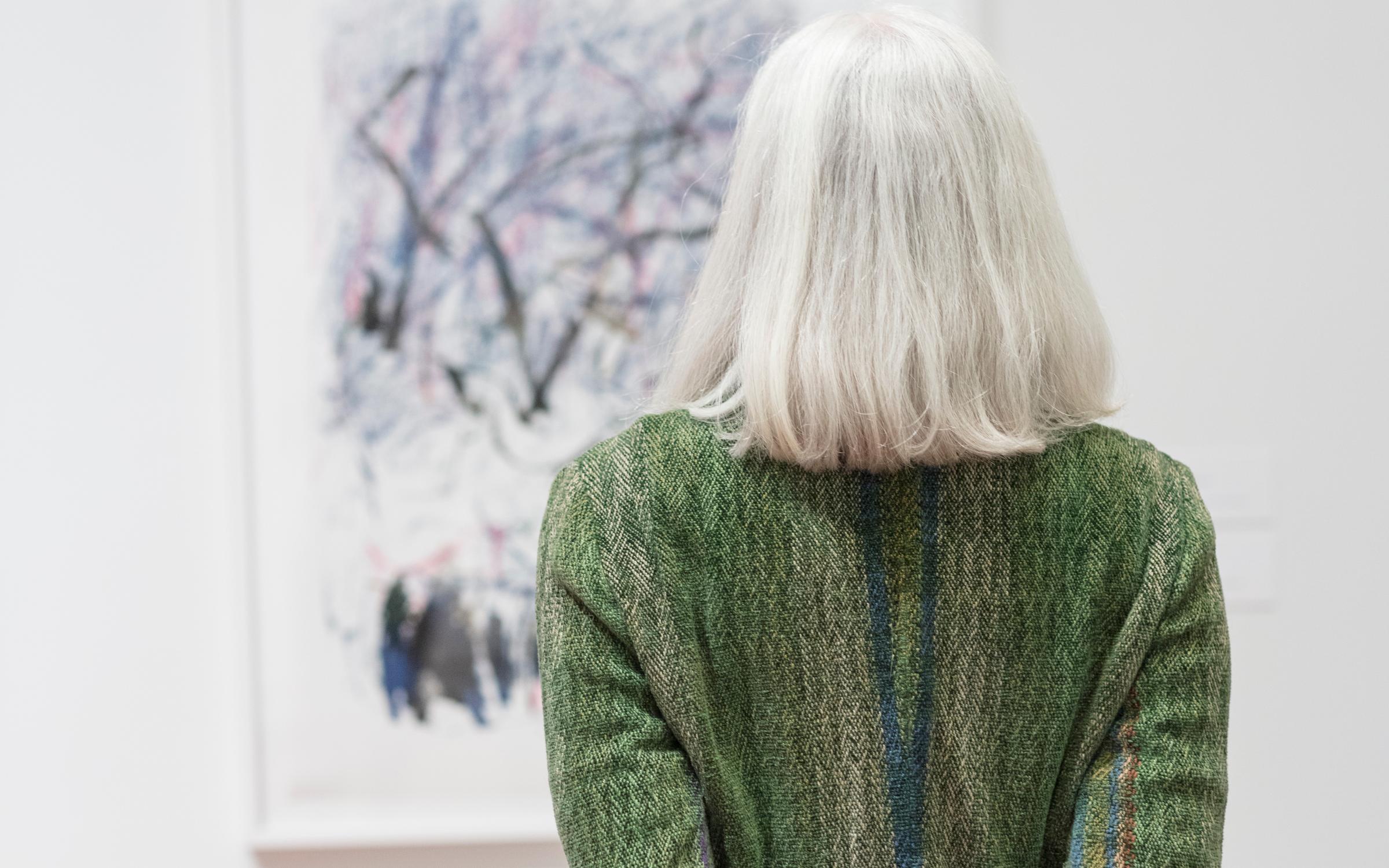 A woman with white hair and a green sweater looking at an abstract painting