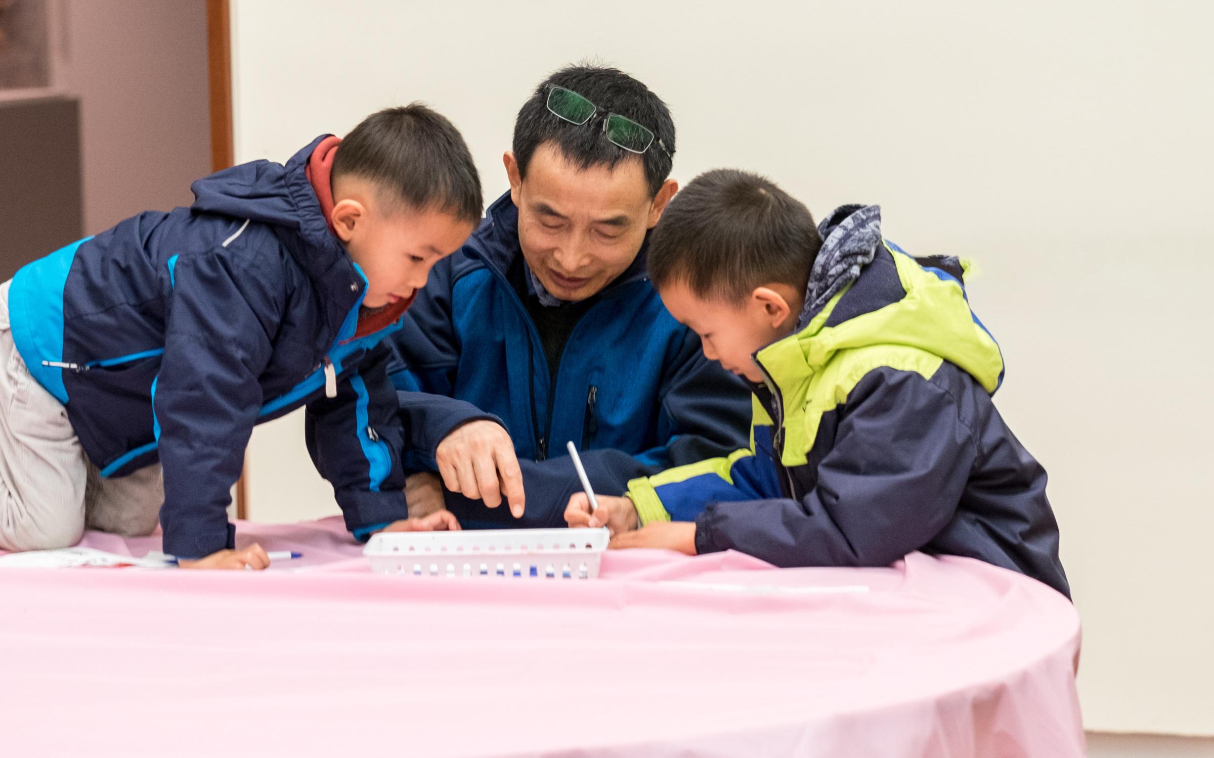 An Asian father and two sons drawing on a table with a pink cloth.