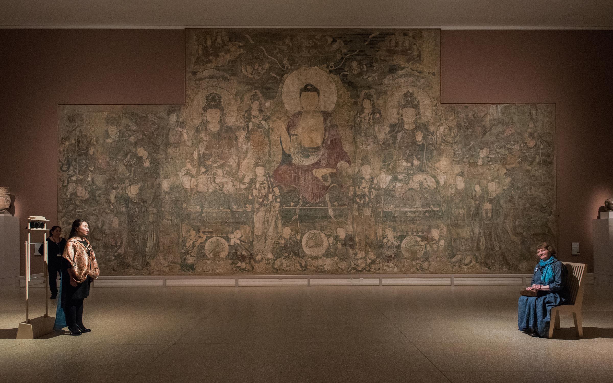 Sonic Blossom at the Met 2015 two people sit in a museum gallery about 20 feet apart. One is a singing to the other as the sit in a wooden chair with a high back.