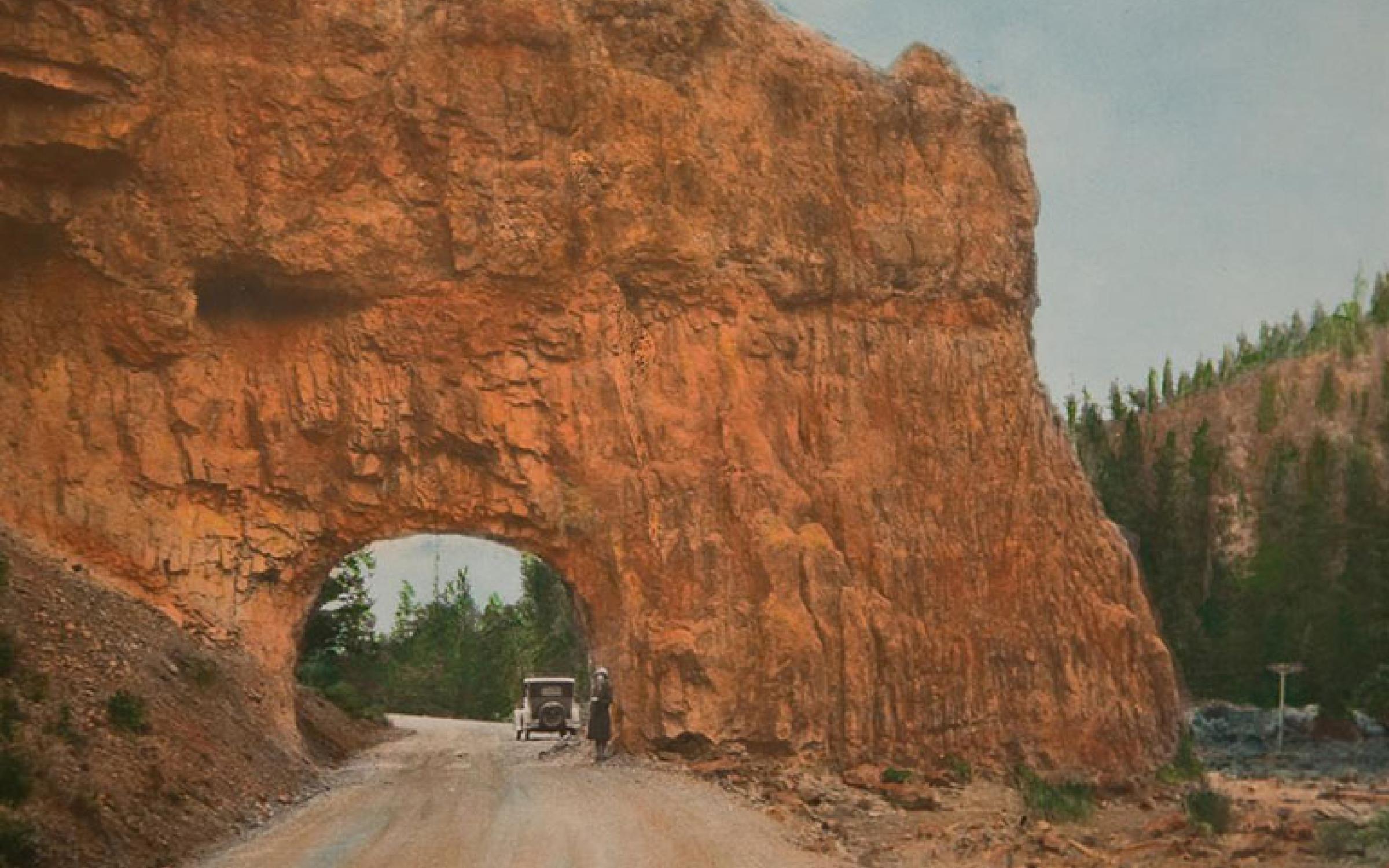 an antique hand colored photograph of a red rock wall wtih a tunnel going through it there is a woman standing next to a car in the tunnel