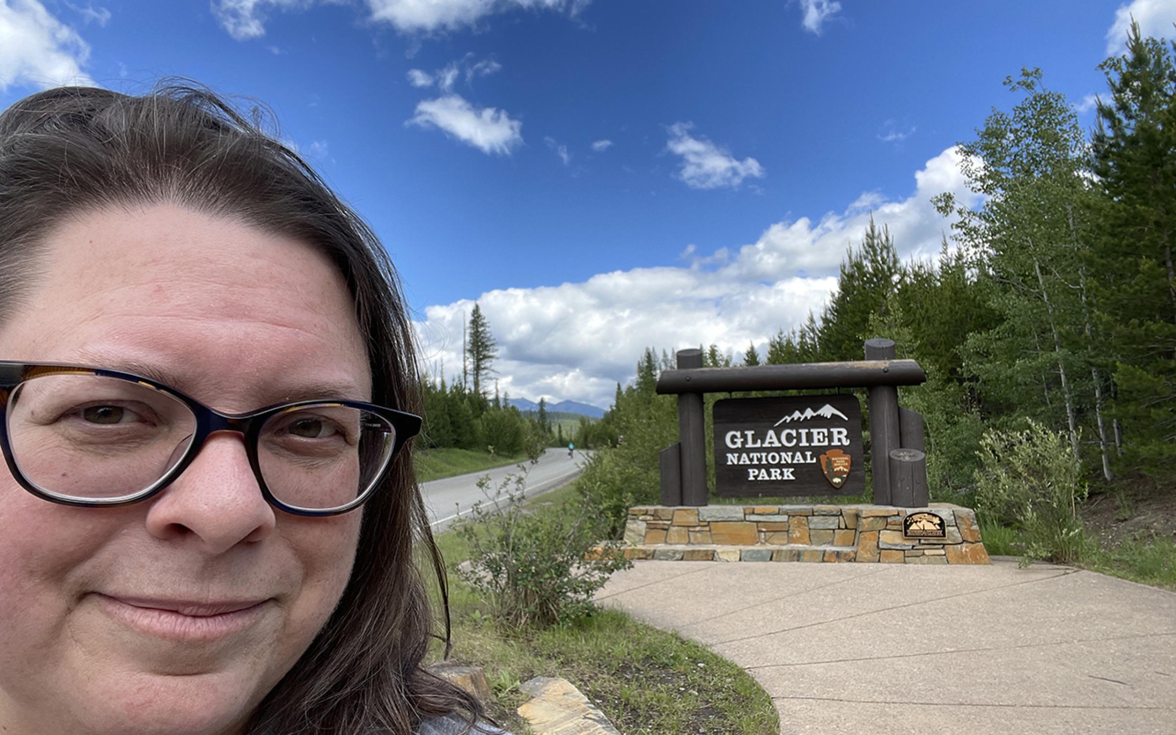 A woman with brown hair and black glasses taking a selfie in front of a national park sign and green grass.
