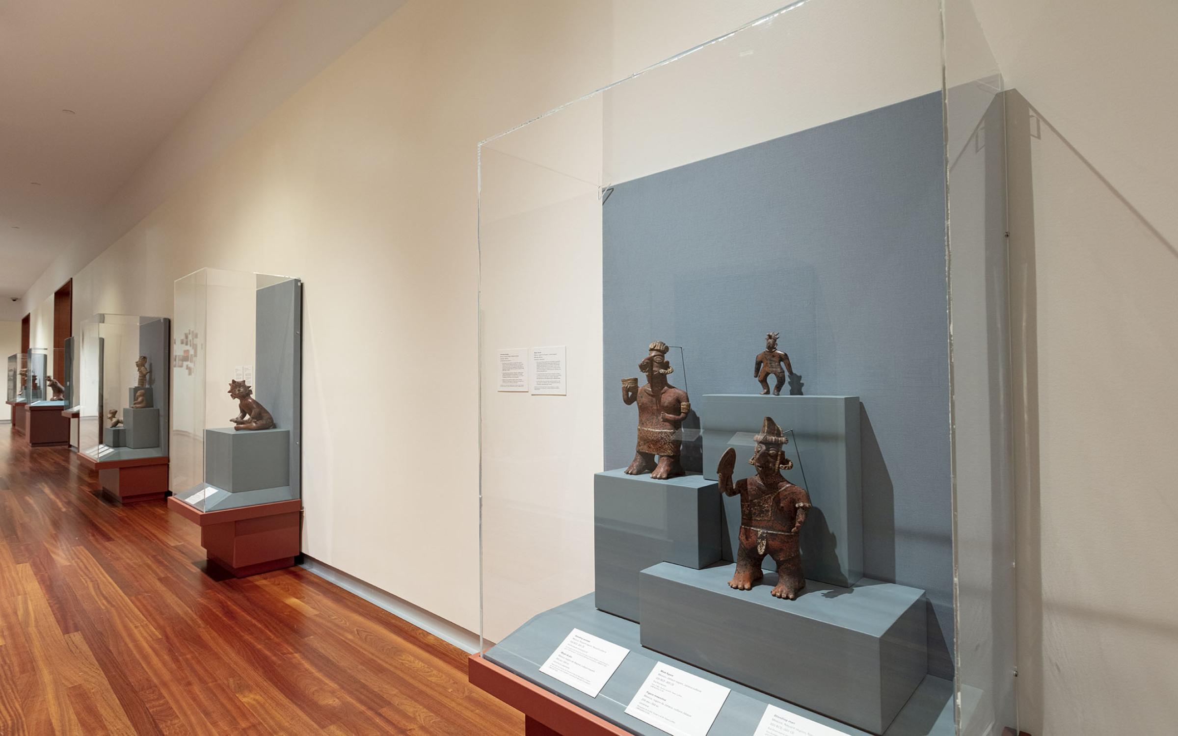 Small, carved figures stand in a glass display case against a white wall. There are multiple similar display cases going down the wall in the background.