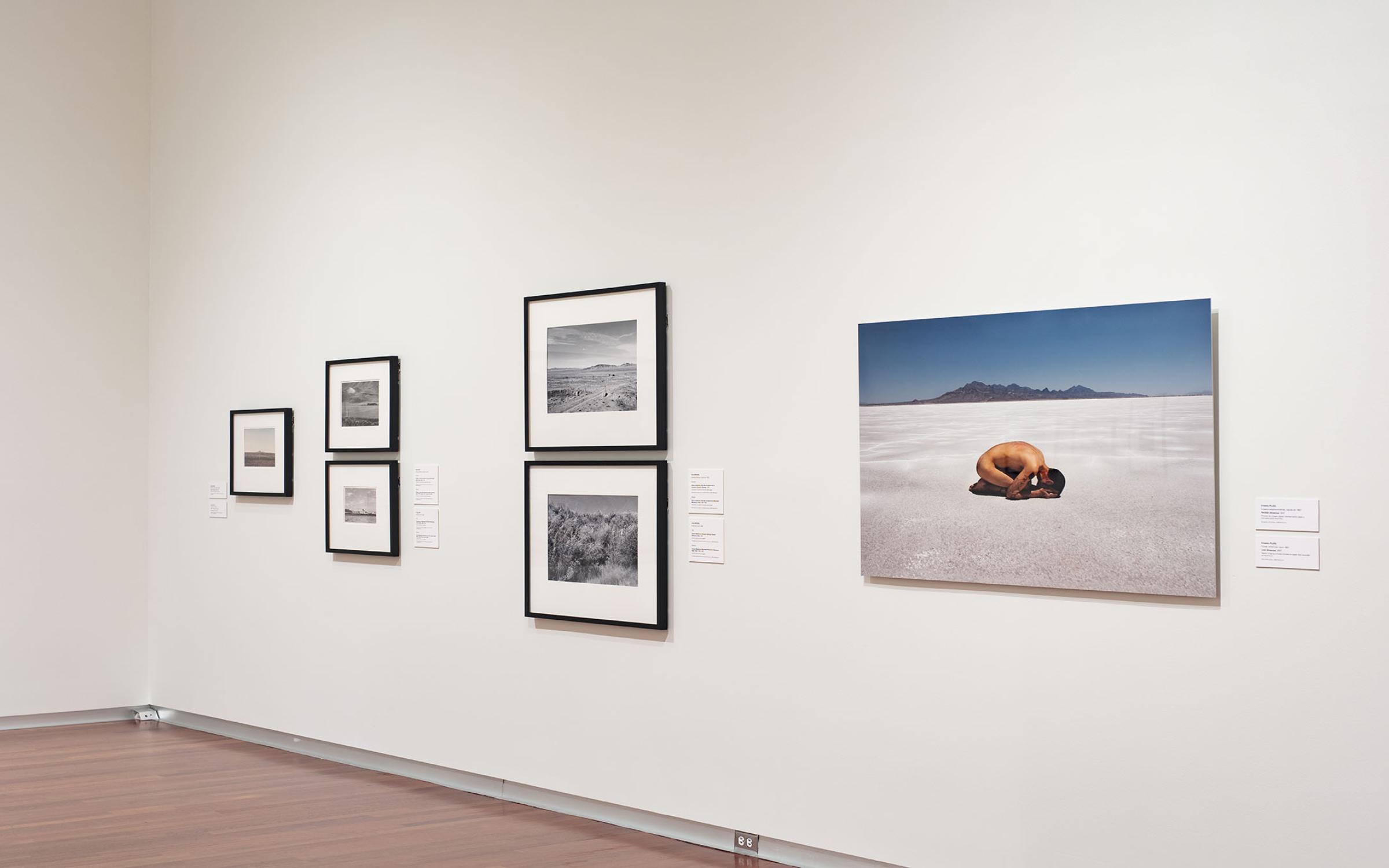 Modern & Contemporary Gallery Framed photos are stacked above one another in two's on a white wall. There's a large photo of a naked person, folded over on the ground next to the framed images. The walls are white.