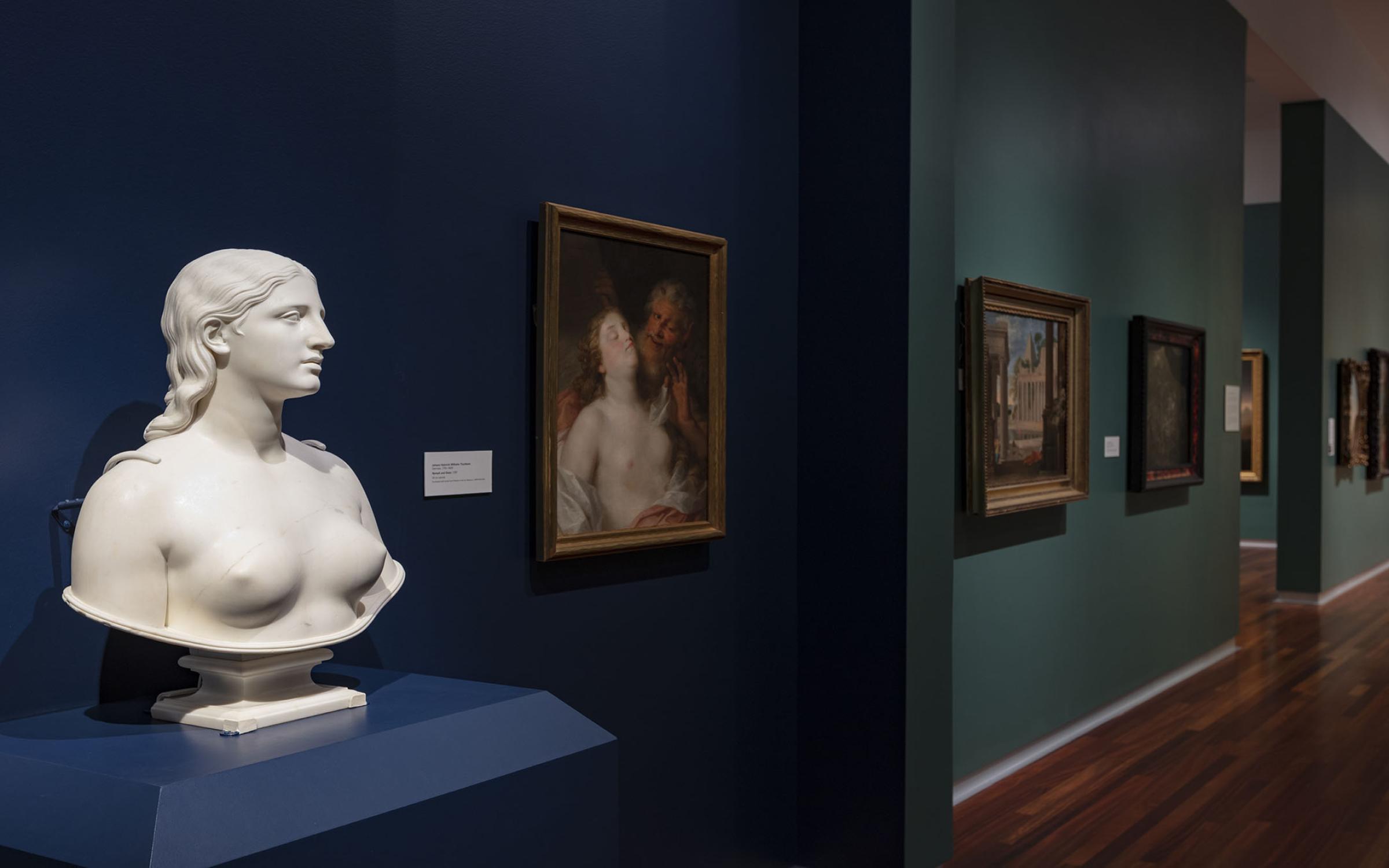 A white statue of a nude woman's bust sits on a dark blue platform. There are paintings on the jewel-toned walls in the background.