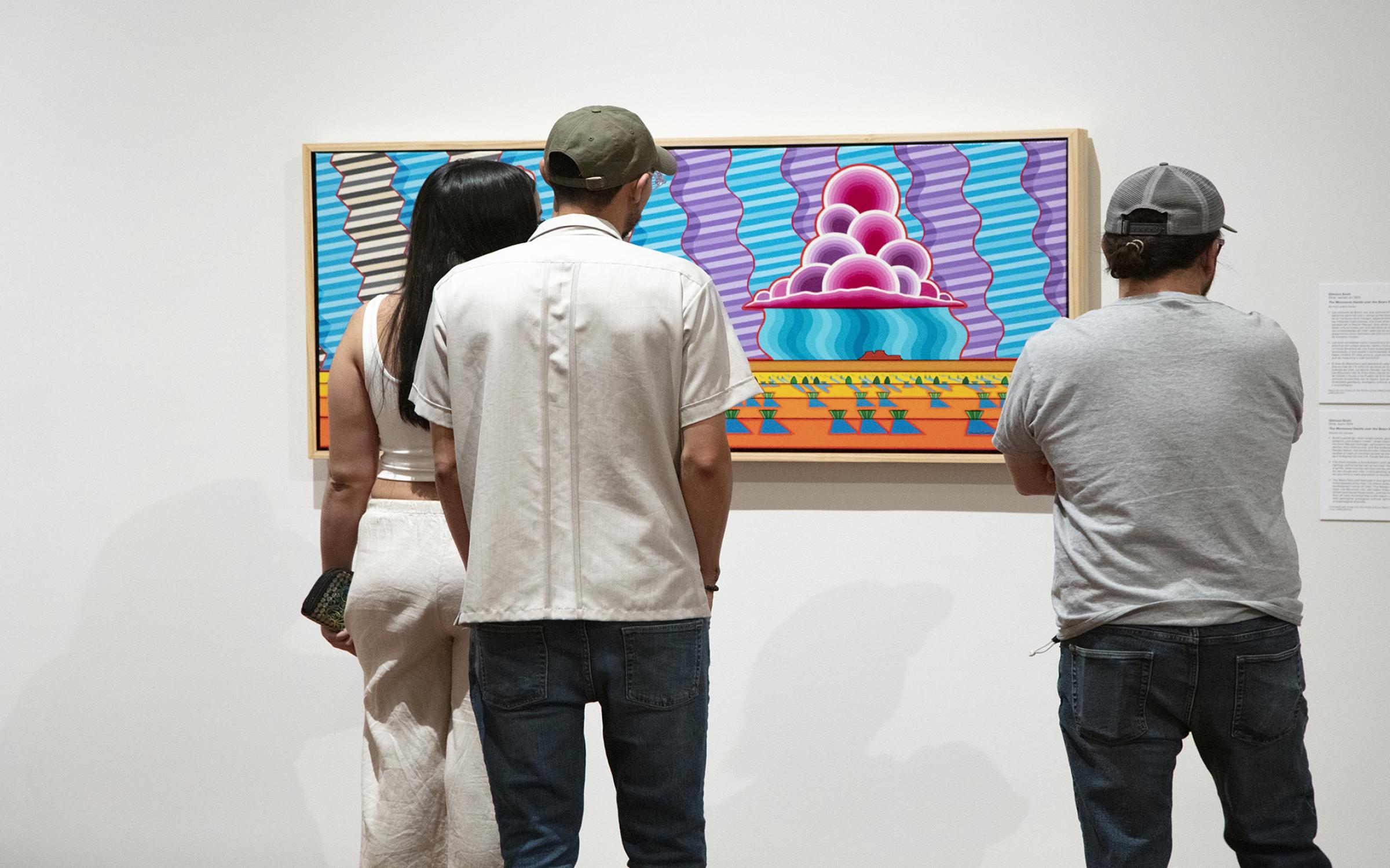 Three people with dark hair, stand with their backs to the camera, looking at bright, colorful artwork on a white wall.