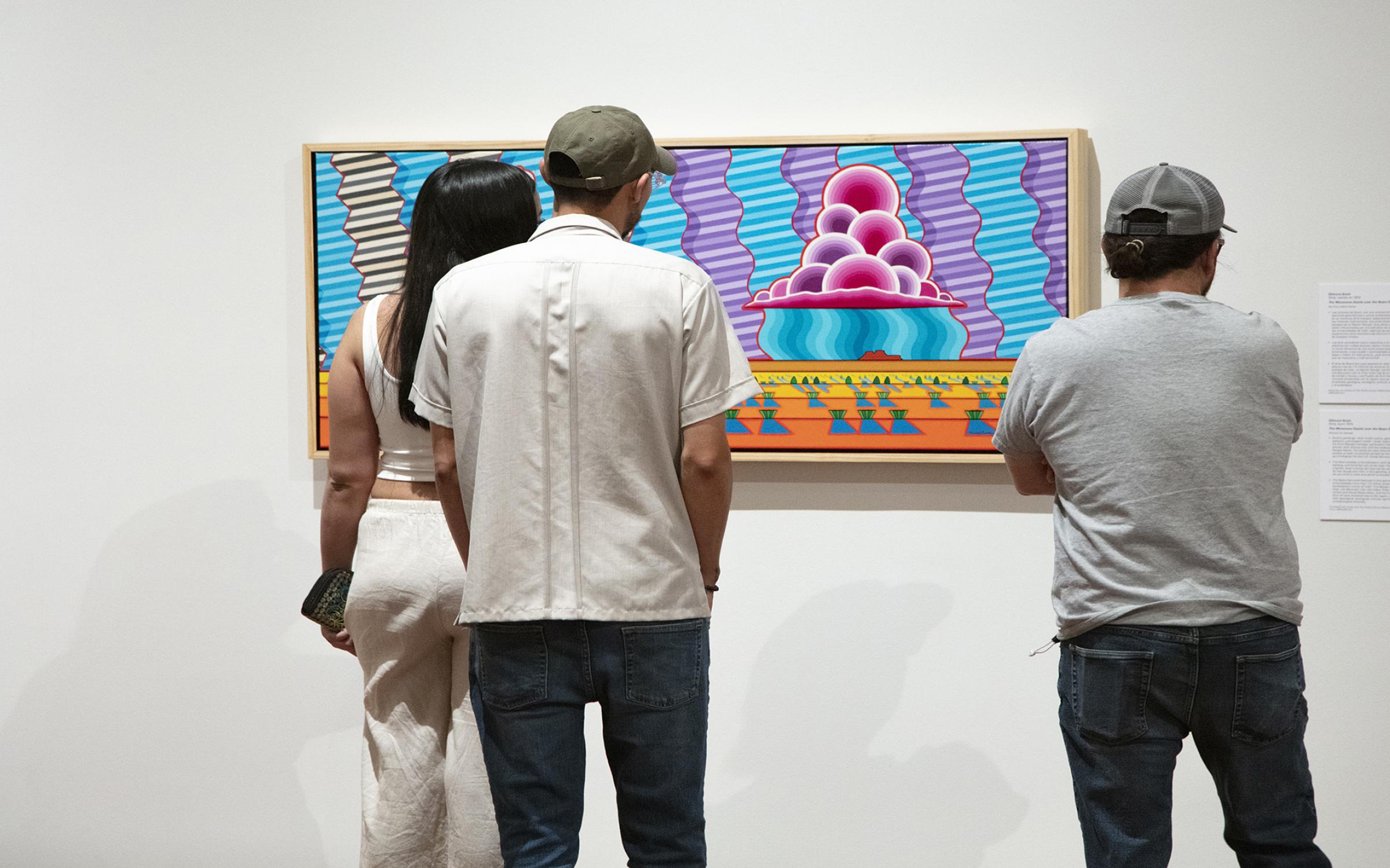 Plan Your Visit Three people with dark hair, stand with their backs to the camera, looking at bright, colorful artwork on a white wall.
