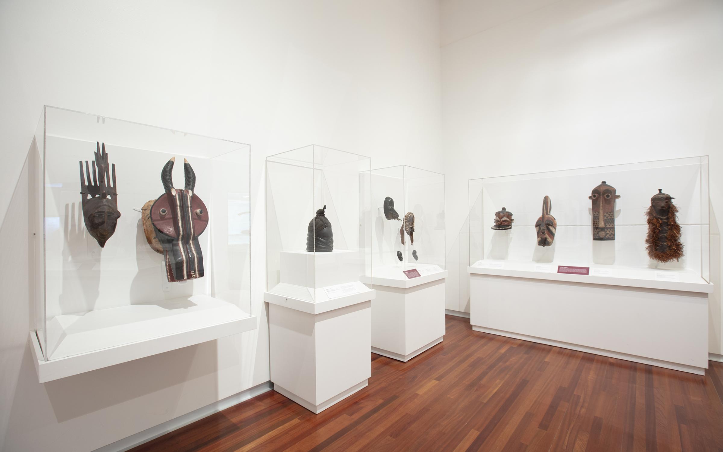 Brown colored masks and figures sit in clear display cases against white walls in a large, open gallery room.