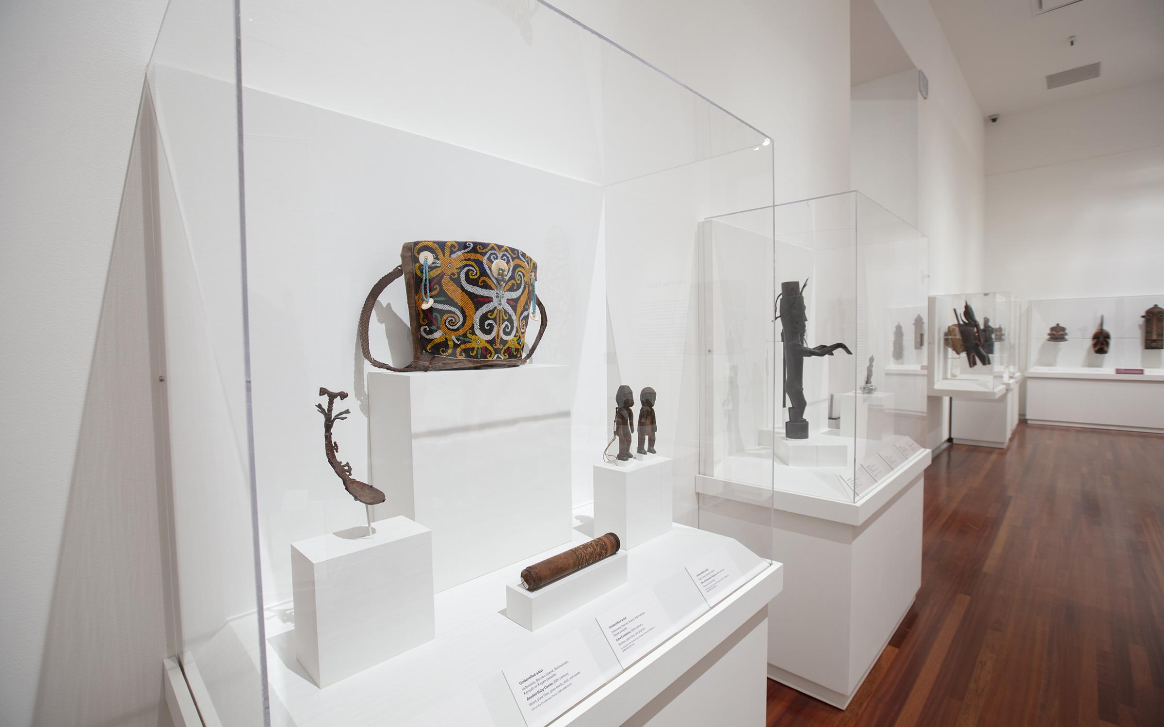 A colorful, beaded basket sits on a white shelf in a clear display case. There are other small, carved items in the same case. There are multiple items in display cases in the background. 