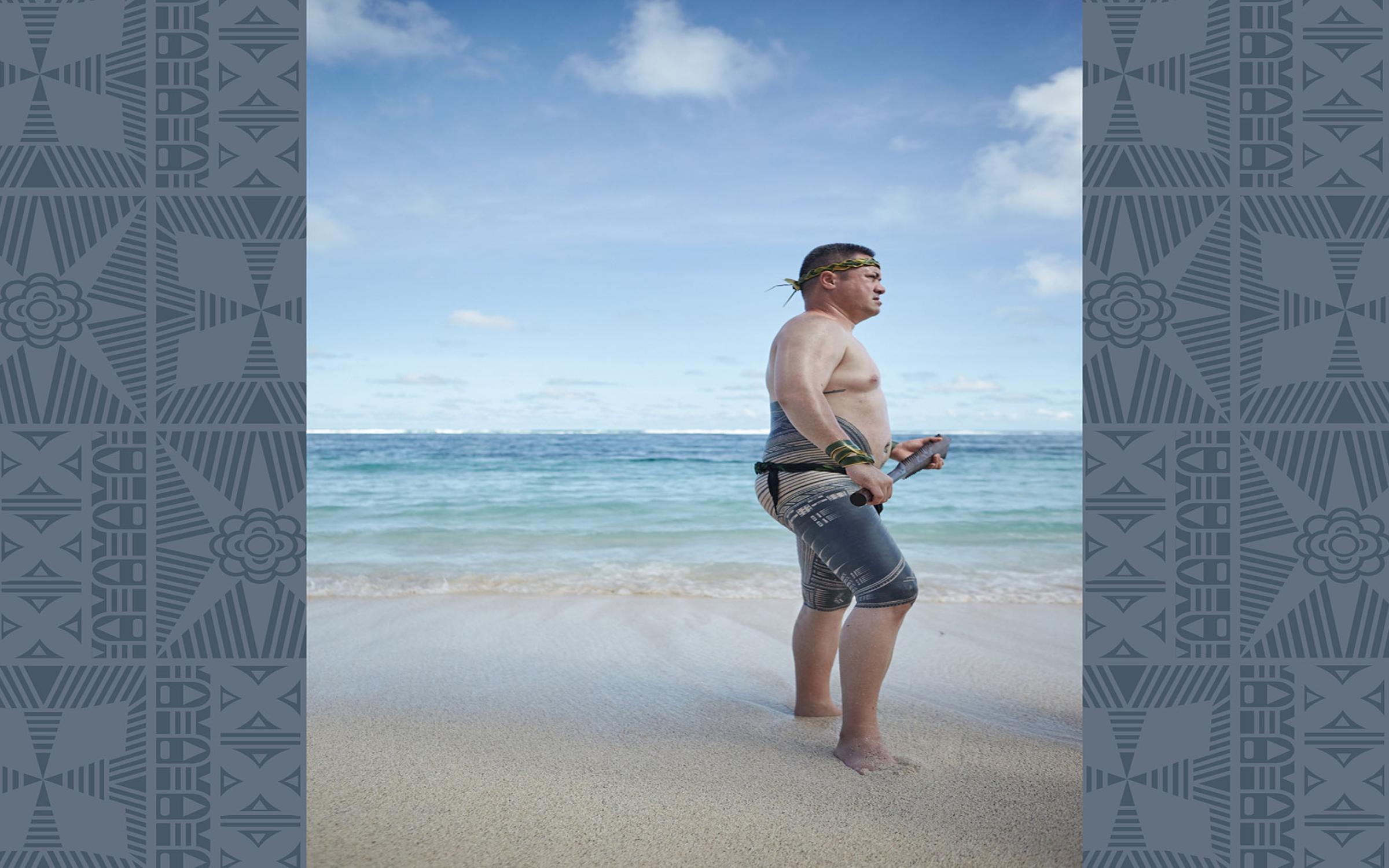 A man wears traditional clothing and no shirt, standing in the sand on a beach. He has dark tattoos on his legs and hips.