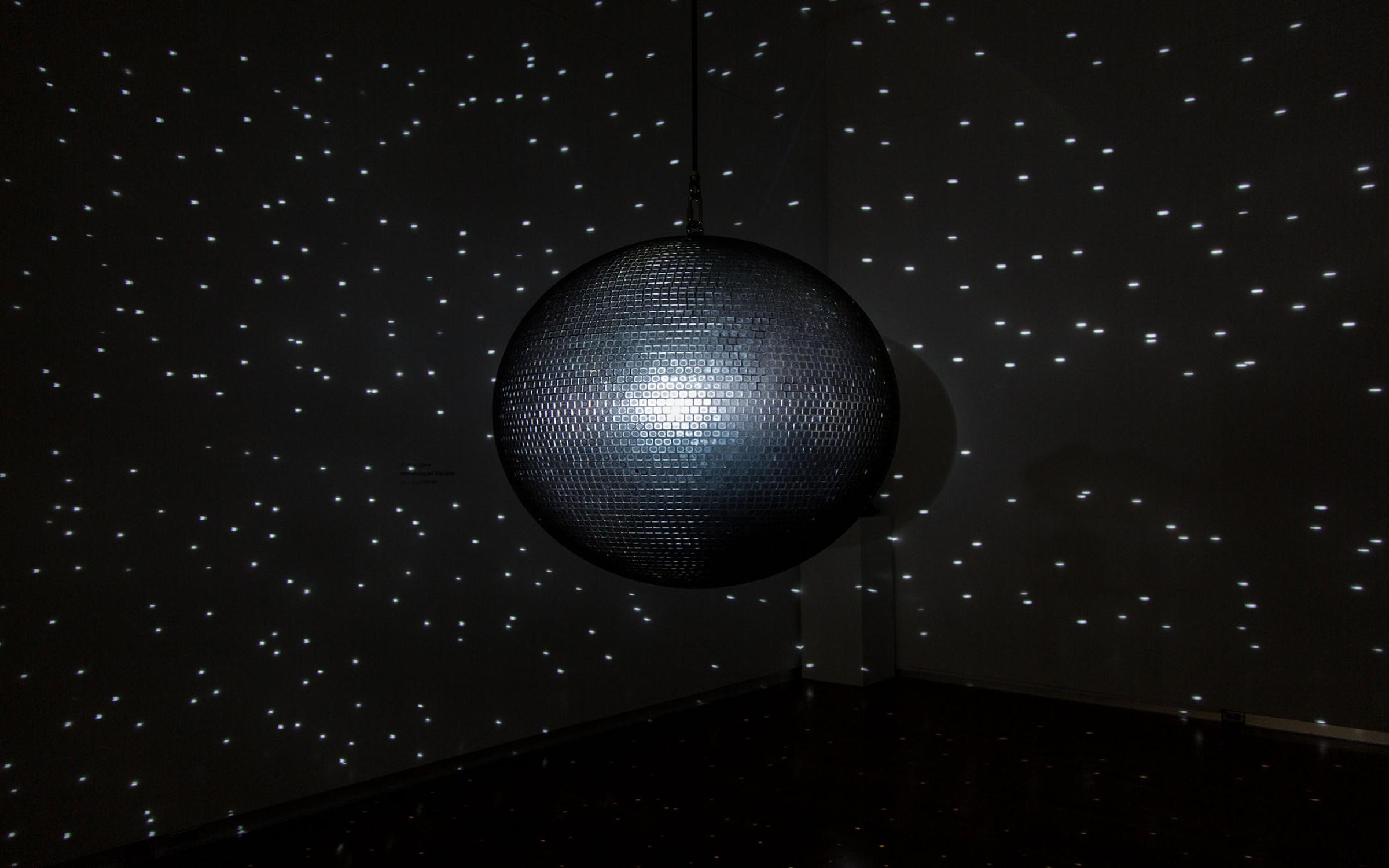 A large, silver disco ball hangs in a dark room, reflecting small dots of bright light on the walls.