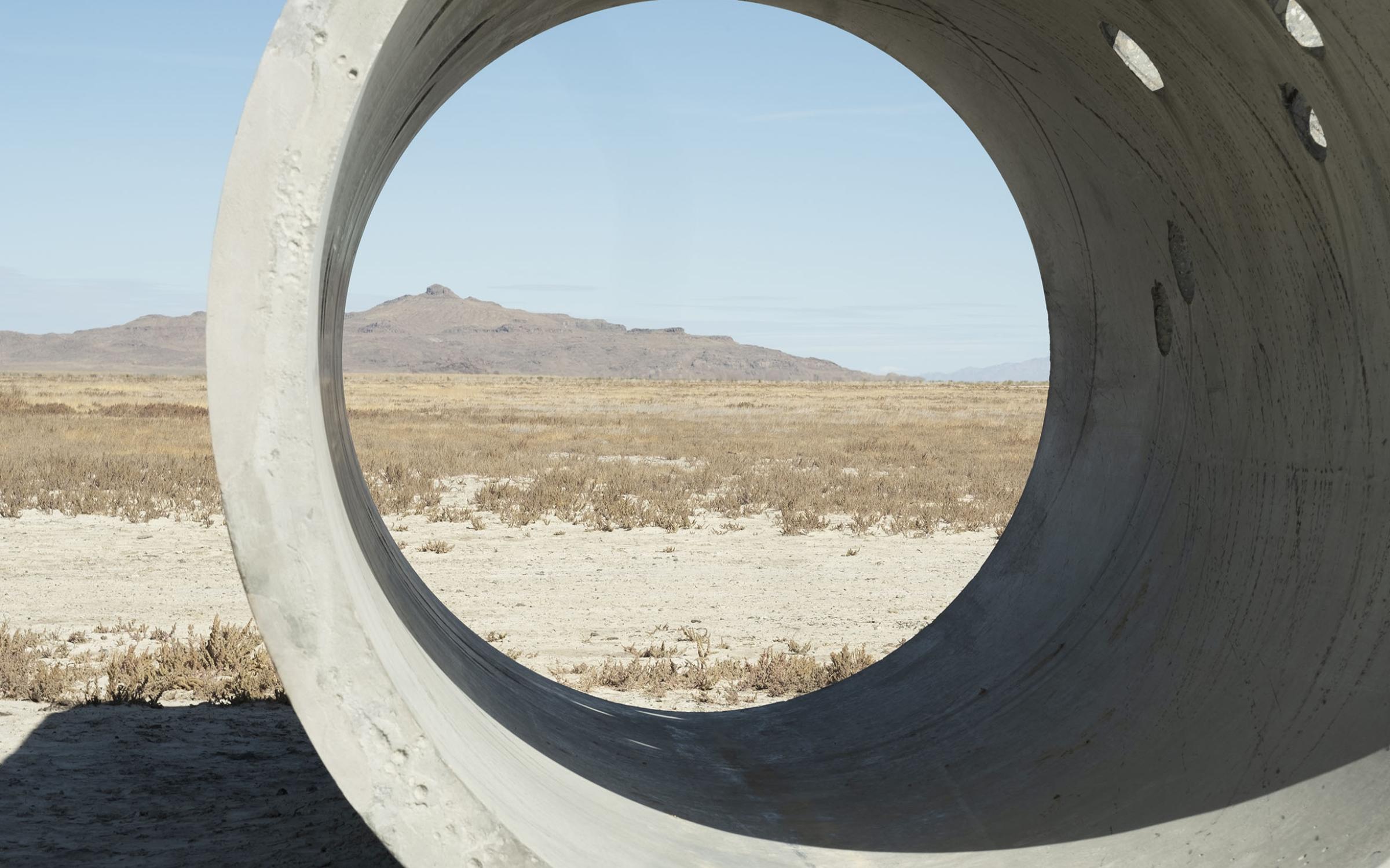 A large concrete cylinder sits in the middle of an open desert landscape. There is tan sand and dry, green plants on the ground. A mountain sits in the background against a light blue sky.