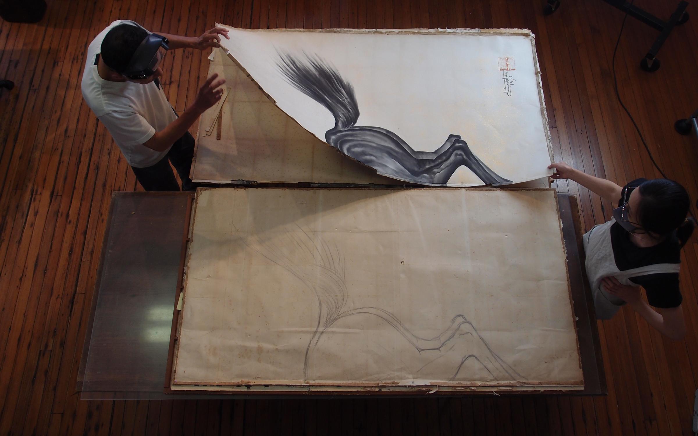 Two people stand on either side of a large table, handling large pieces of paper with a painting of the back end of a black horse.