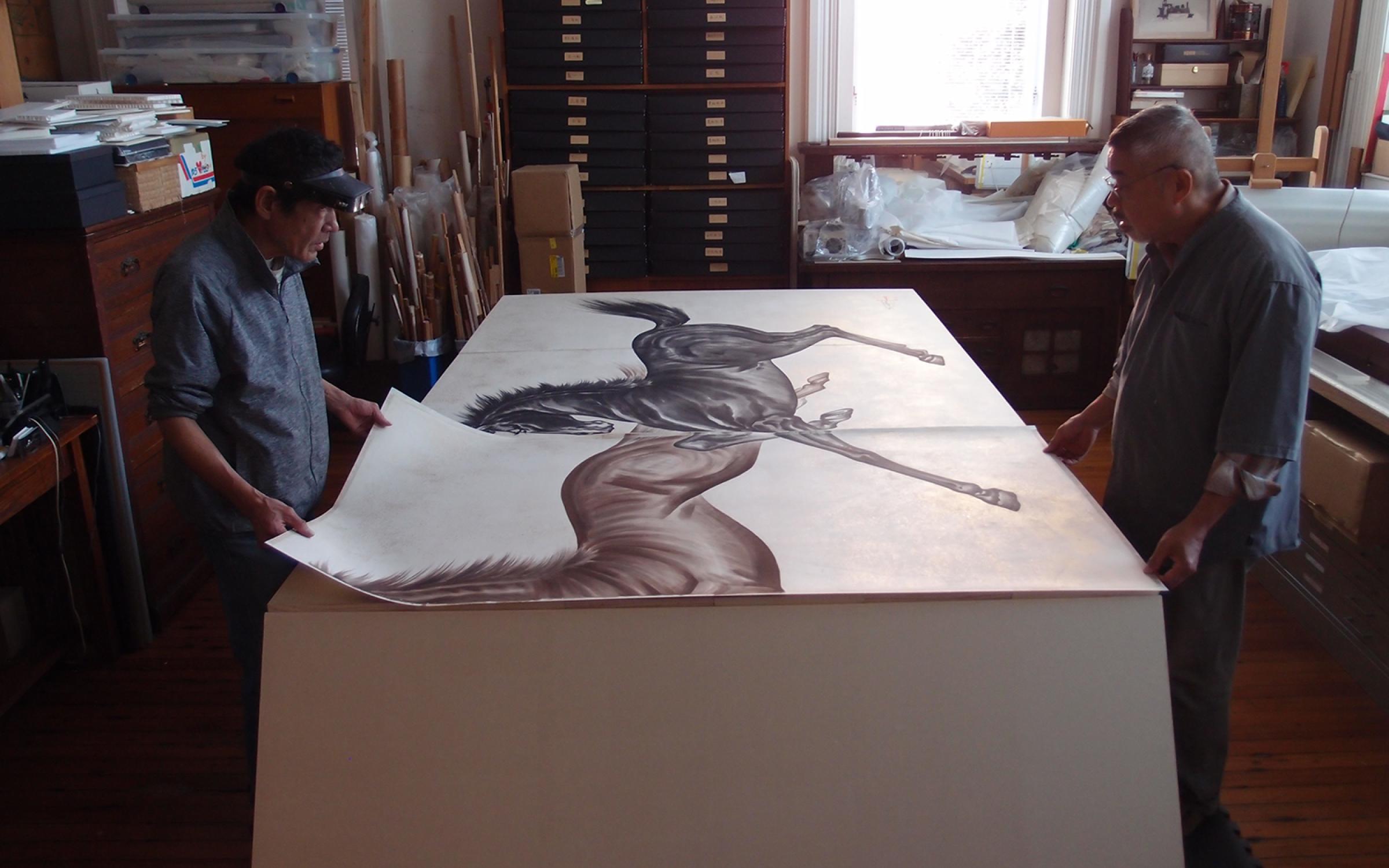 A photo of two men standing on either side of a large table, touching paper with paintings of two horses.