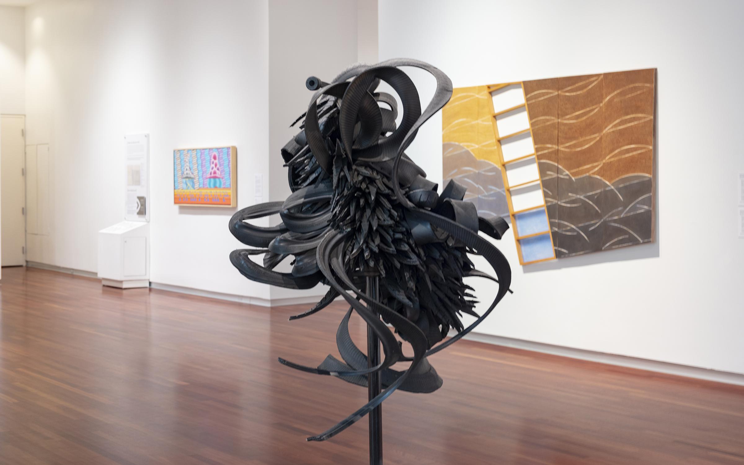 A sculpture made of shreds of car tires stands in the middle of a large gallery room. There are colorful paintings hung on the white walls in the background.