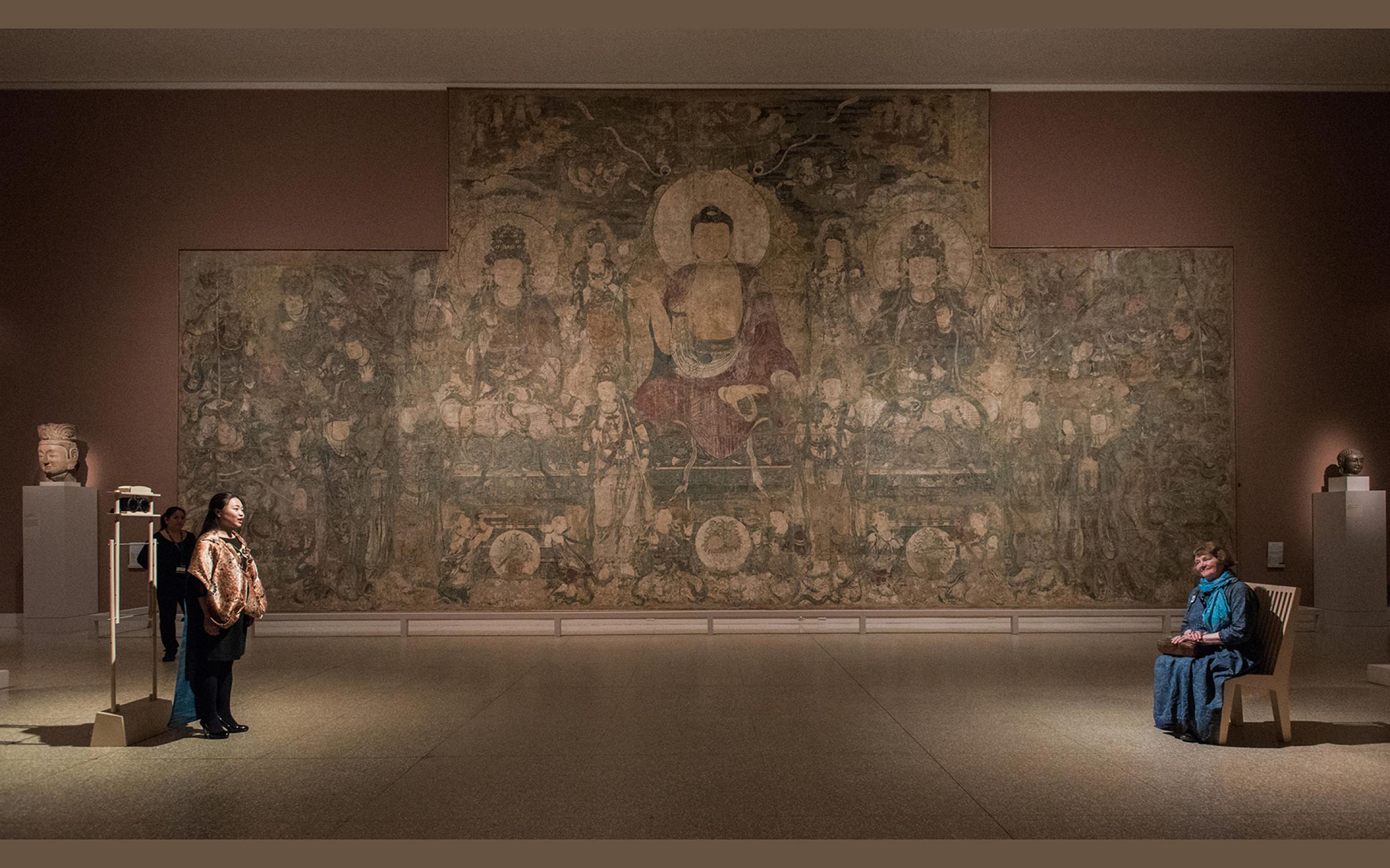 A photograph of two people on opposites sides of a large room; one is sitting in a blue dress, and the other is standing and singing. There's a large mural with eastern deities in the background.