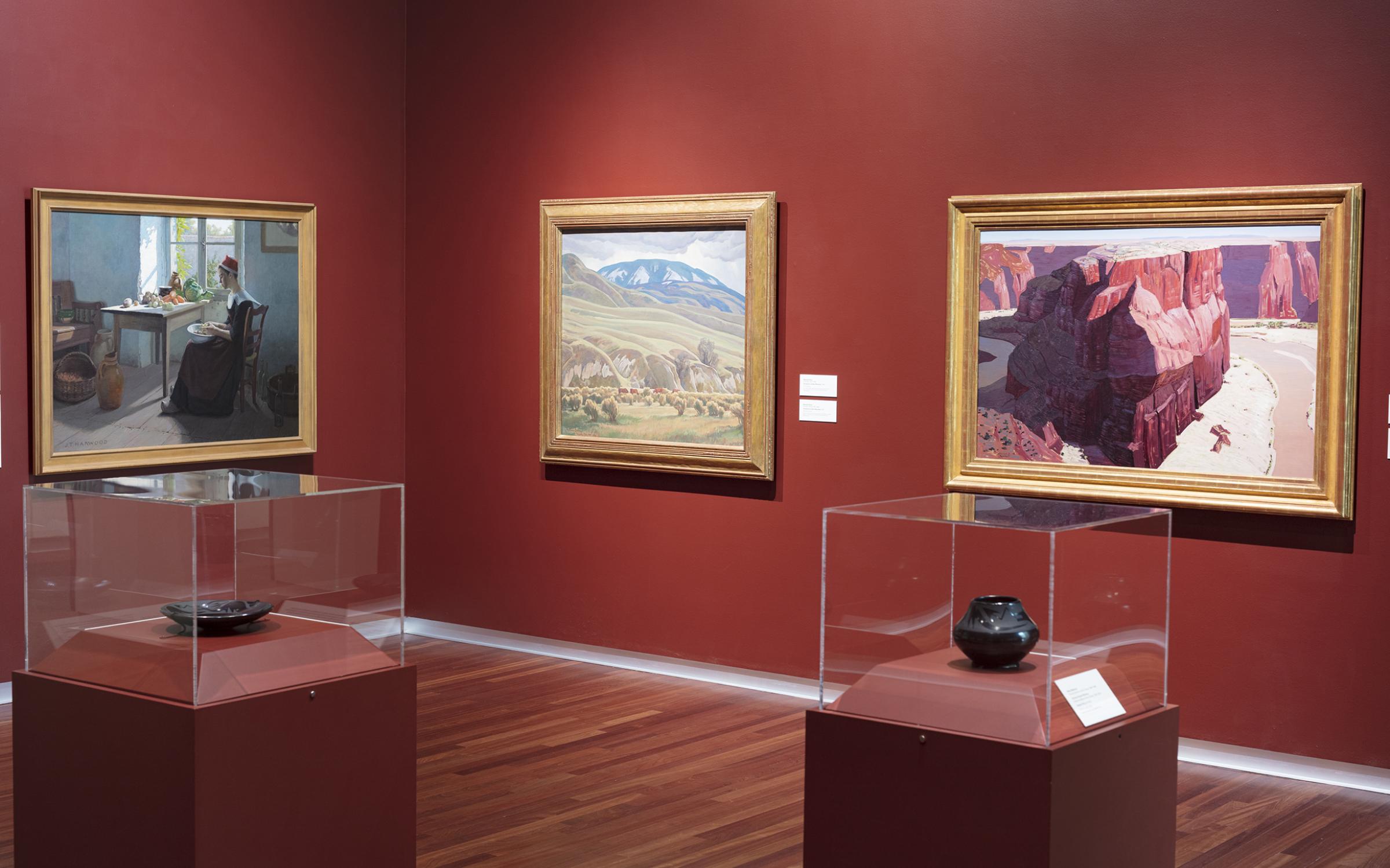 Three framed paintings line the burgundy-colored walls in the background. There are two dark pieces of pottery in separate glass display cases on similarly colored stands in the middle of the room.
