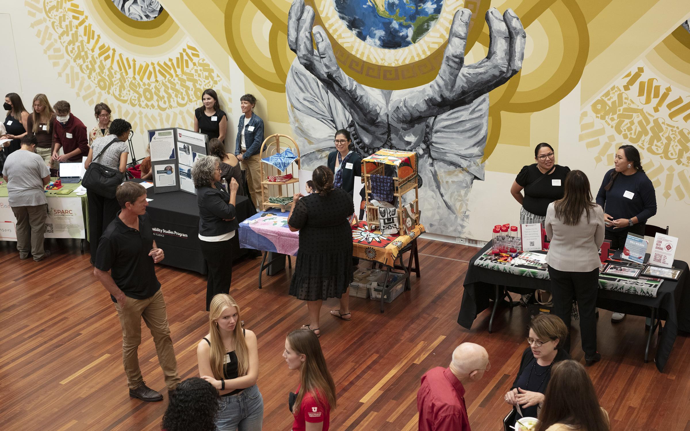 A large group of people stand around a large room, talking in small groups. There are tables near the far wall with different materials and goods that people are looking at. There is a mural in the background of two hands holding and earth in front of a golden mandala shape.