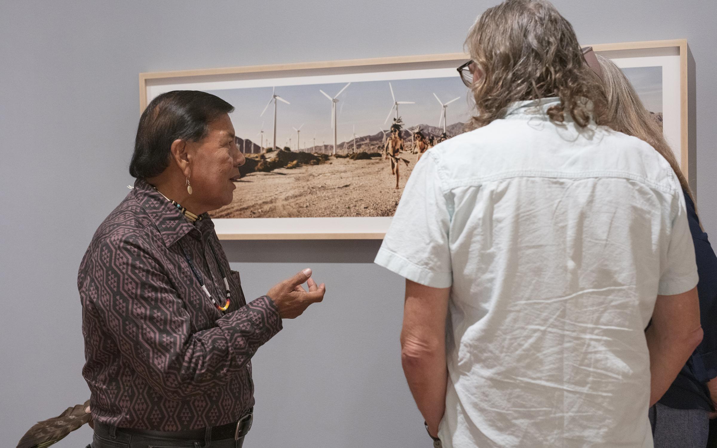 A native man in a button up shirt with dark hair speaks to two white people with grey hair. They are all looking at a framed artwork on a light grey wall.
