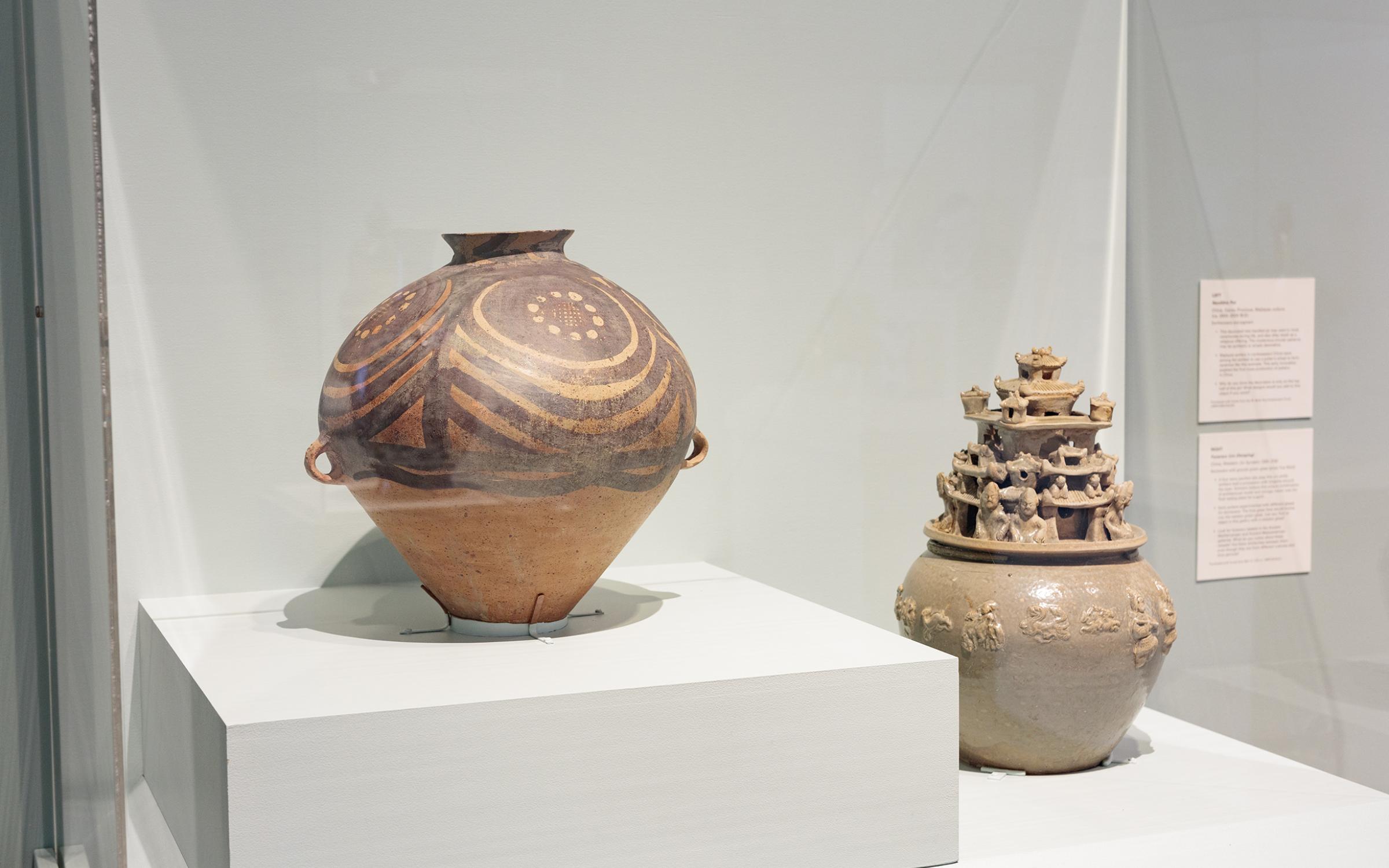 Two large pieces of carved and painted pottery sit inside a glass display case on a mint-colored stand.