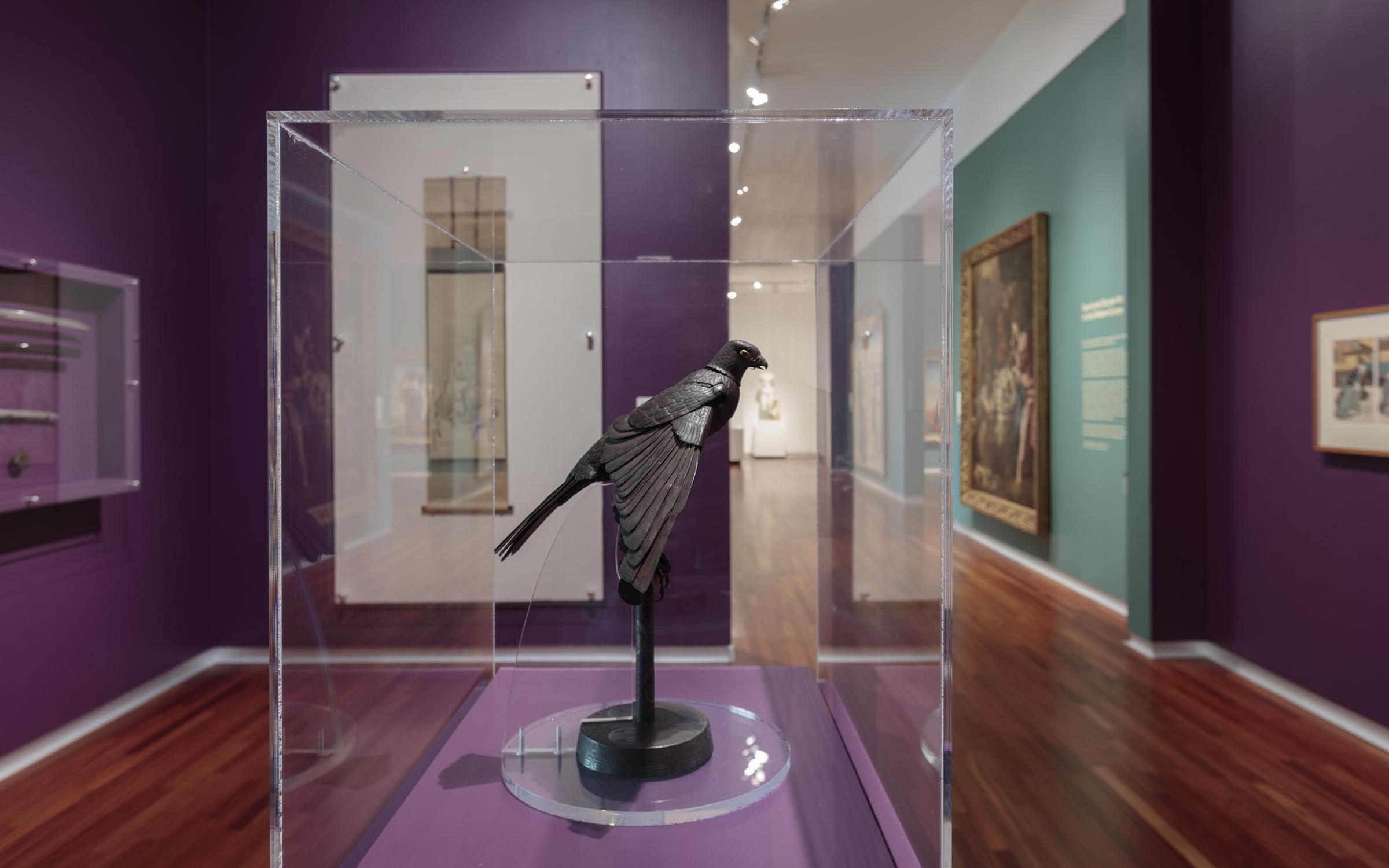 A brown sculpture of a small bird sits on a narrow stand in a glass case in the center of the room. The walls are a deep purple and there are samurai swords hanging on the wall to the left in a glass display case. There is a long painted scroll on the purple wall in the background and dark green walls with framed art down the hallway to the right.